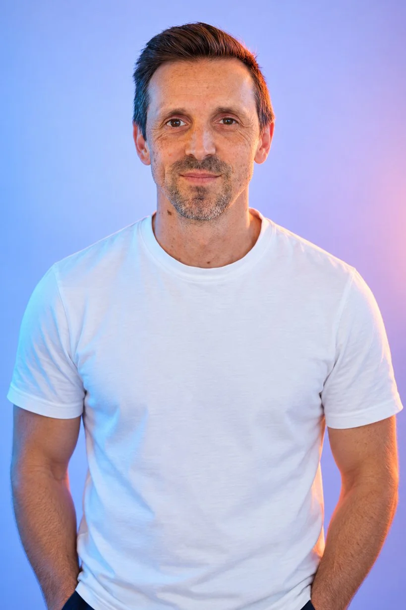 A middle-aged man with short brown hair and a beard, wearing a plain white t-shirt, standing against a gradient background of purple, blue, and pink colors.