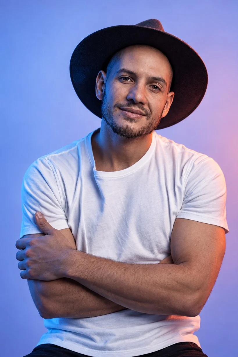 A young man with a beard and mustache wearing a black wide-brimmed hat and a white t-shirt, standing with arms crossed against a blue and purple gradient background.