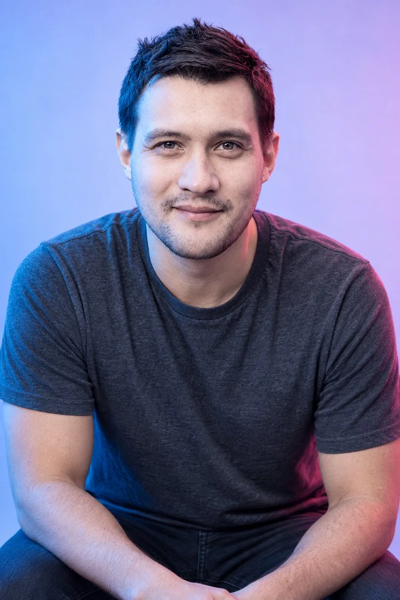 A young man with short brown hair and a light beard, smiling, wearing a dark gray T-shirt, sitting against a gradient background of blue and pink lighting.