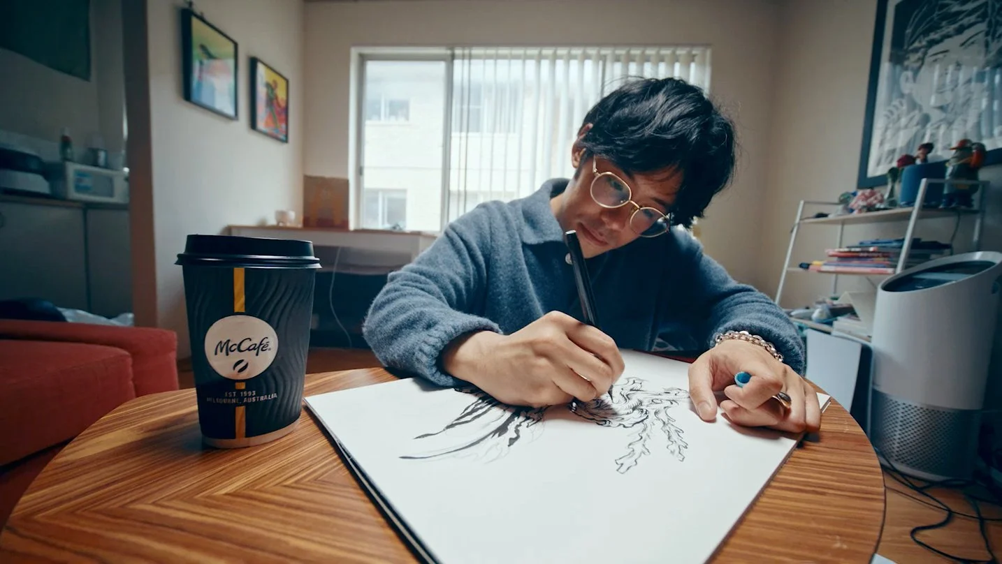 A woman drawing a lion on a large sheet of paper at a wooden table in a cozy home office, with a McCafe coffee cup nearby.