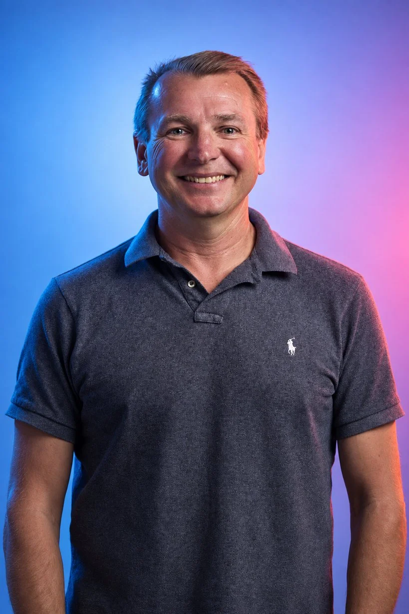Portrait of a smiling man with short blonde hair, wearing a dark gray polo shirt, against a colorful gradient background of blue and pink hues.
