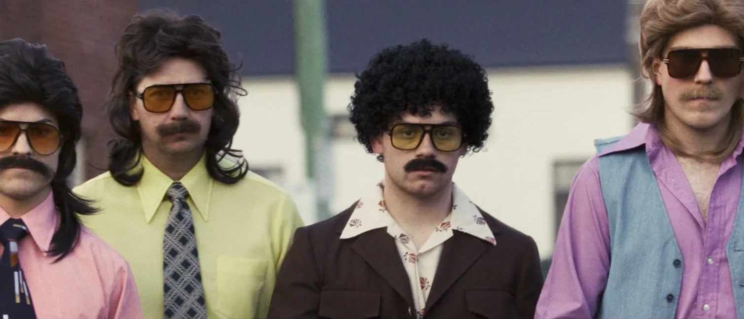 Four men with mustaches and sunglasses standing outdoors, wearing colorful vintage clothing from the 1970s.
