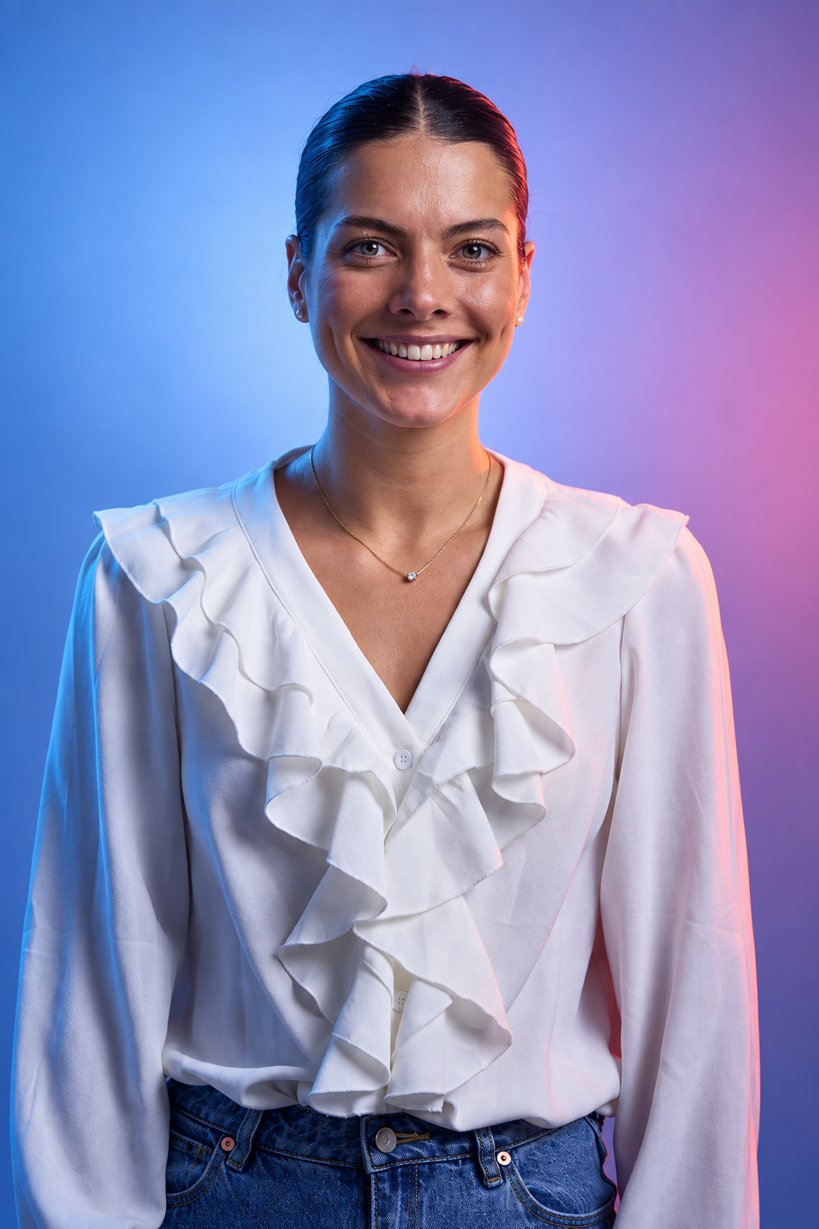 A smiling woman with dark hair tied back, wearing a white blouse with ruffles and blue jeans, standing against a colorful blue and pink gradient background.
