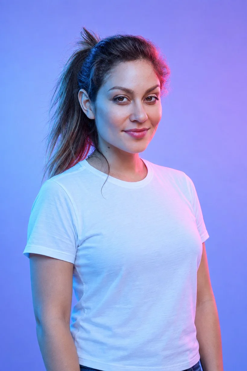 A woman with brown hair tied in a messy ponytail, wearing a plain white T-shirt, standing against a gradient background of blue and purple lighting, smiling slightly.