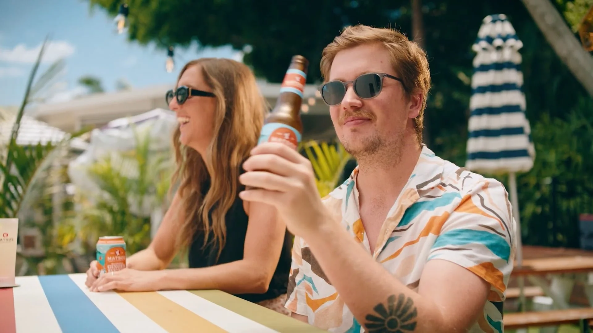 A man with sunglasses and short hair, wearing a colorful short-sleeve shirt, holding a beer bottle, sitting at an outdoor table with a woman, who is also wearing sunglasses and smiling. The scene is set outdoors under a tree with a striped umbrella i