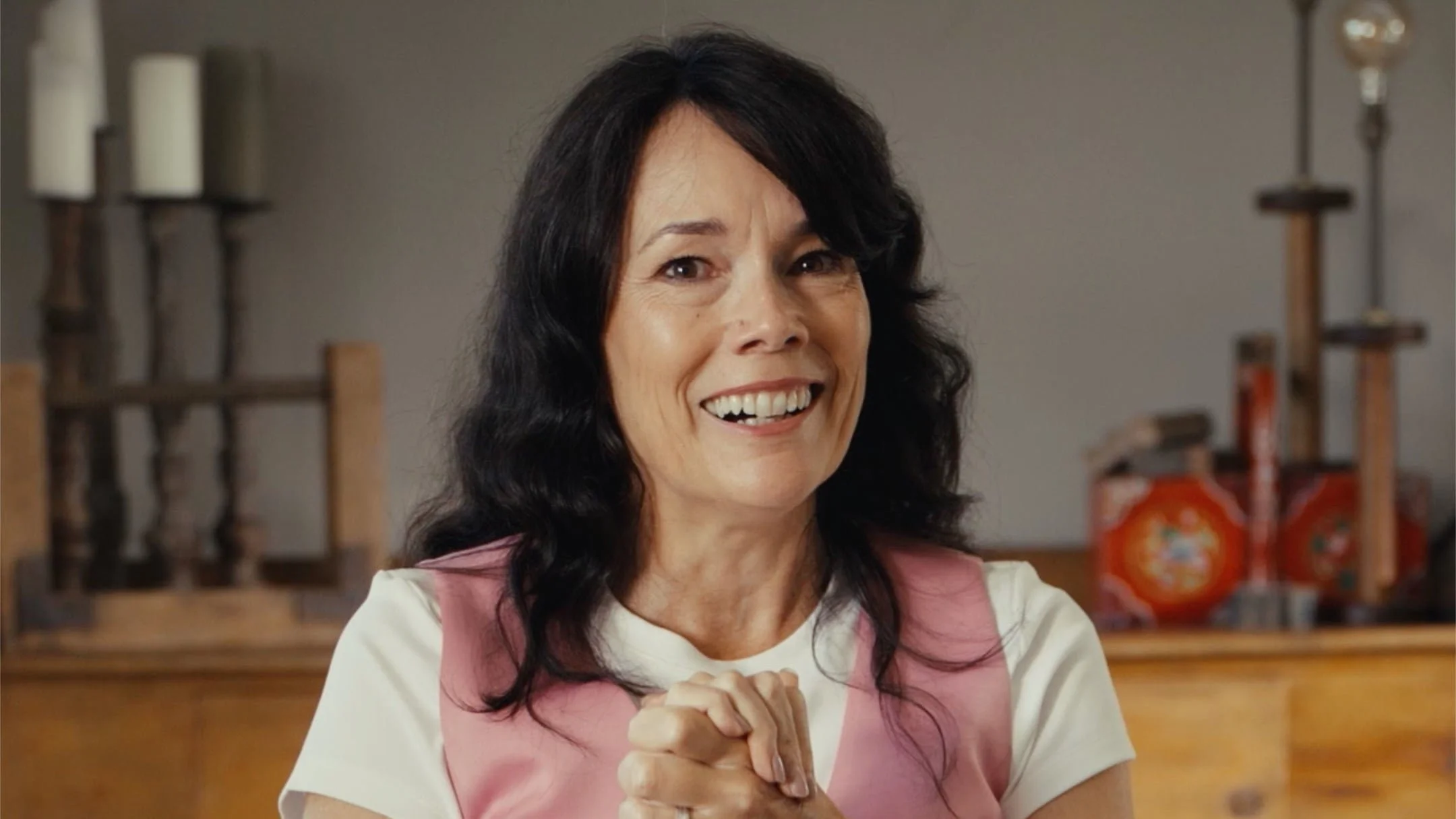 Smiling woman with dark hair in a cheerful setting, with blurred shelves and decorative items in the background.