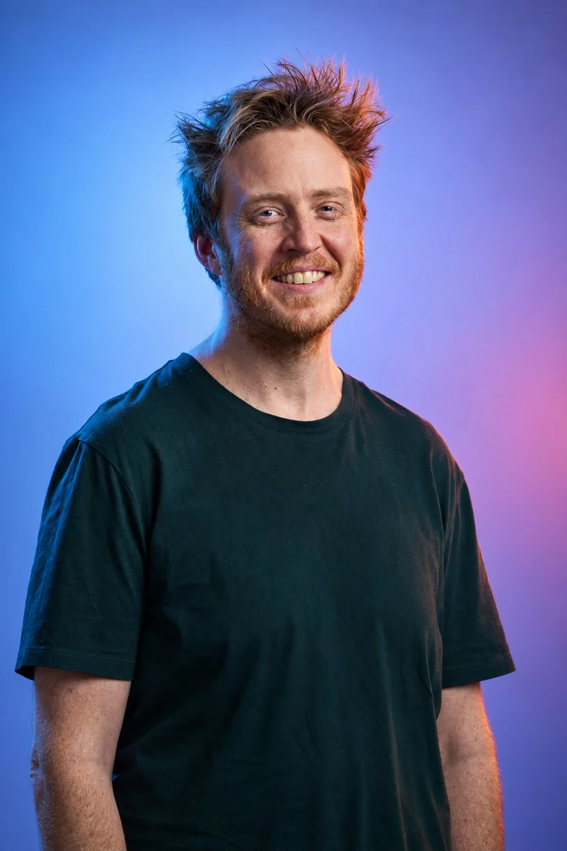A young man with messy red hair and a beard smiling at the camera, wearing a black t-shirt against a colorful gradient background.