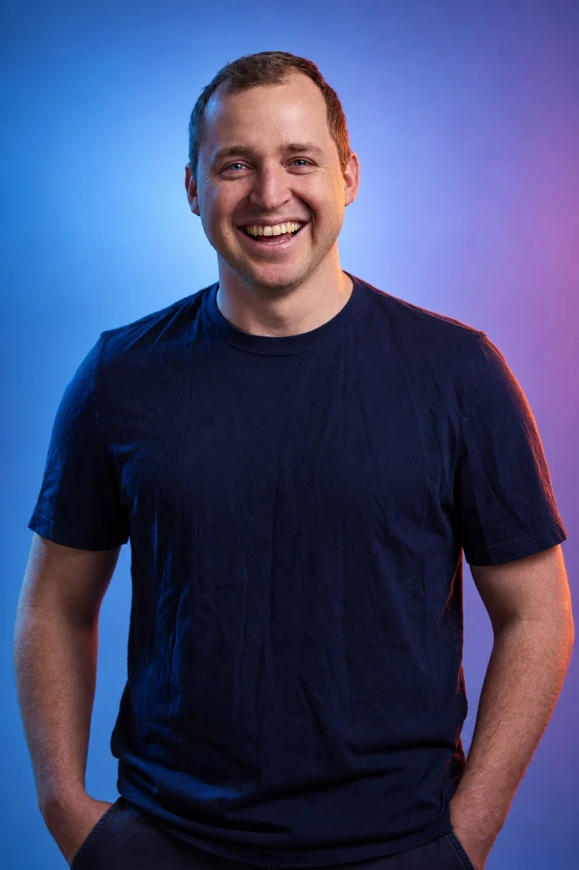 Young man with short hair wearing a navy t-shirt smiling and standing with both hands in his pockets against a blue and purple gradient background.