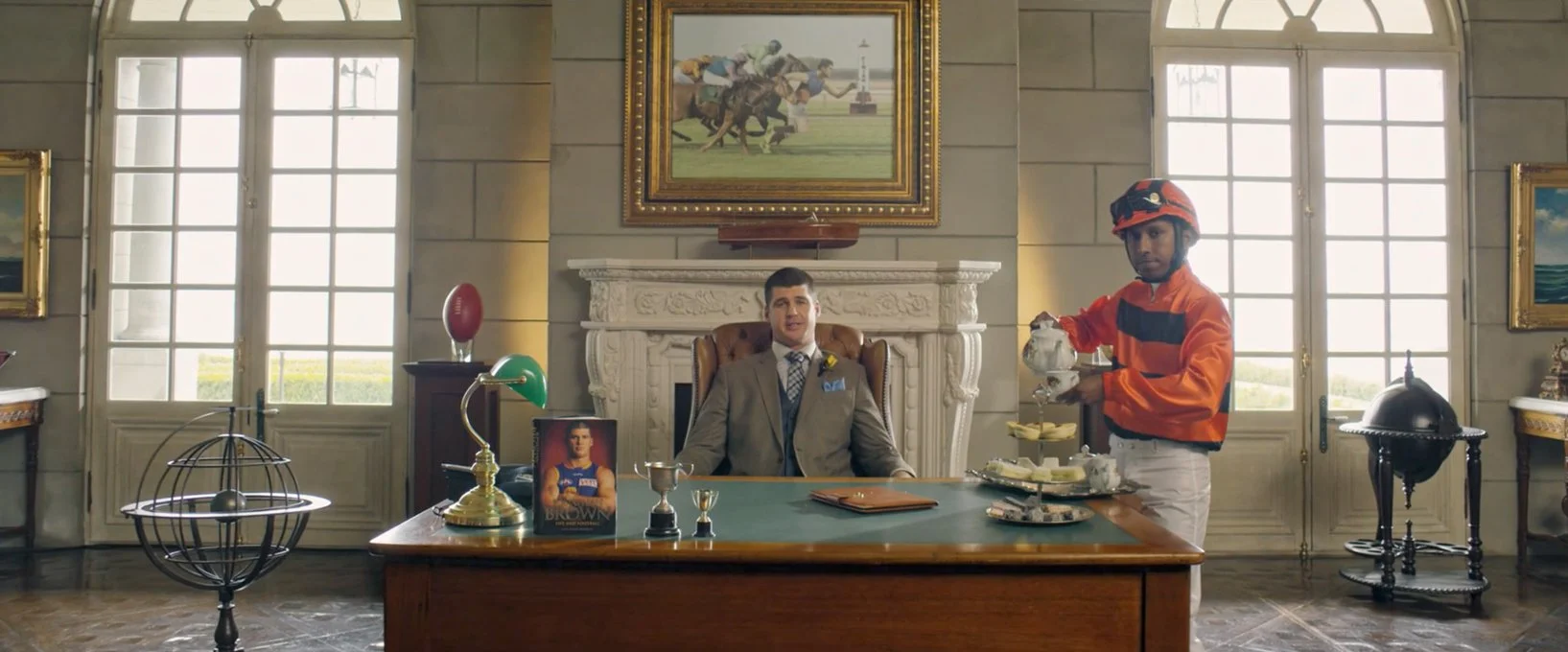 A man in a suit sitting at a large desk in an ornate office with two large windows behind, a fireplace, and framed paintings. A model of an armillary sphere is on the left side of the desk, and a tea set on a stand is on the right, with a jockey in racing gear serving tea.
