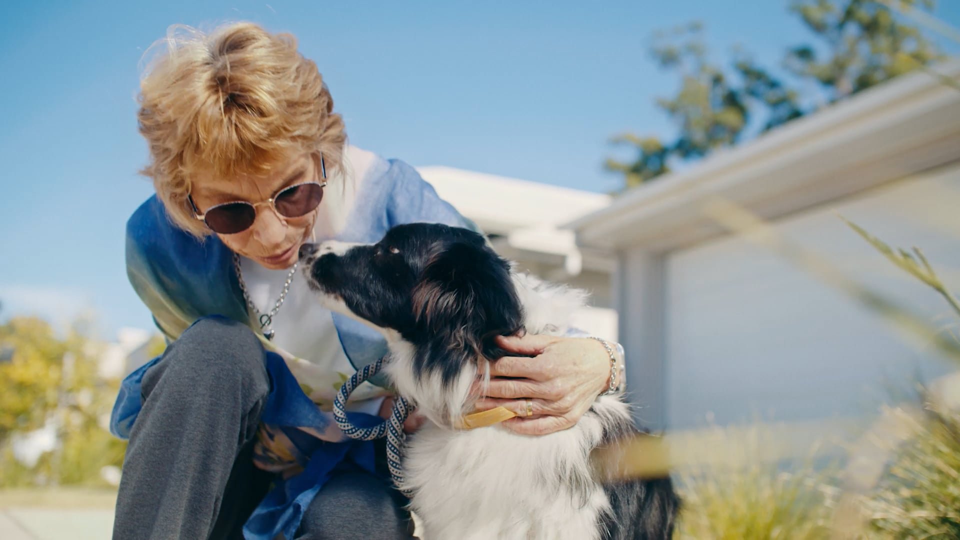An older woman with blonde hair and sunglasses is leaning in to kiss a black and white dog outside on a sunny day, with a house and trees in the background.