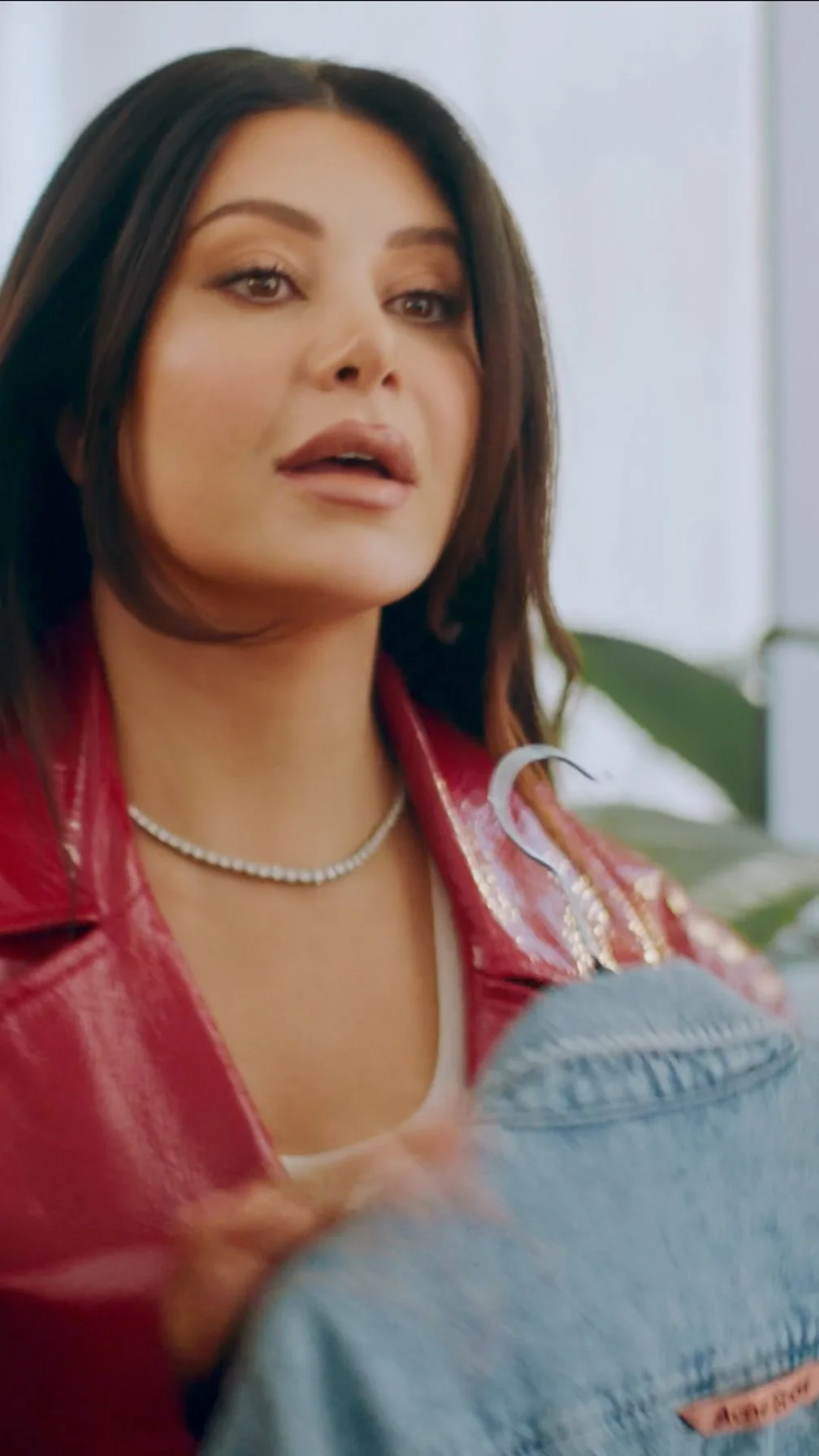 Close-up of a woman with dark hair, wearing a red leather jacket, pearl necklace, and makeup, talking to someone in an indoor setting.