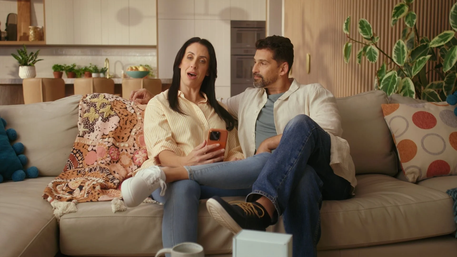 A woman and a man sitting on a beige sofa in a living room, looking at a smartphone together. The woman has long black hair, wears a yellow striped shirt, and white sneakers. The man has short dark hair, a beard, and wears a light-colored shirt and j