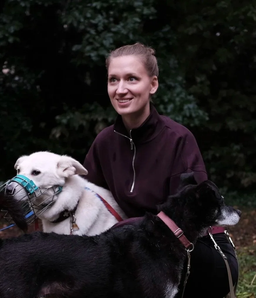 Eine Frau sitzt im Freien mit zwei Hunden, einem weißen Hund mit Maulkorb und einem schwarzen Hund, vor einem Hintergrund aus dunklen Bäumen.
