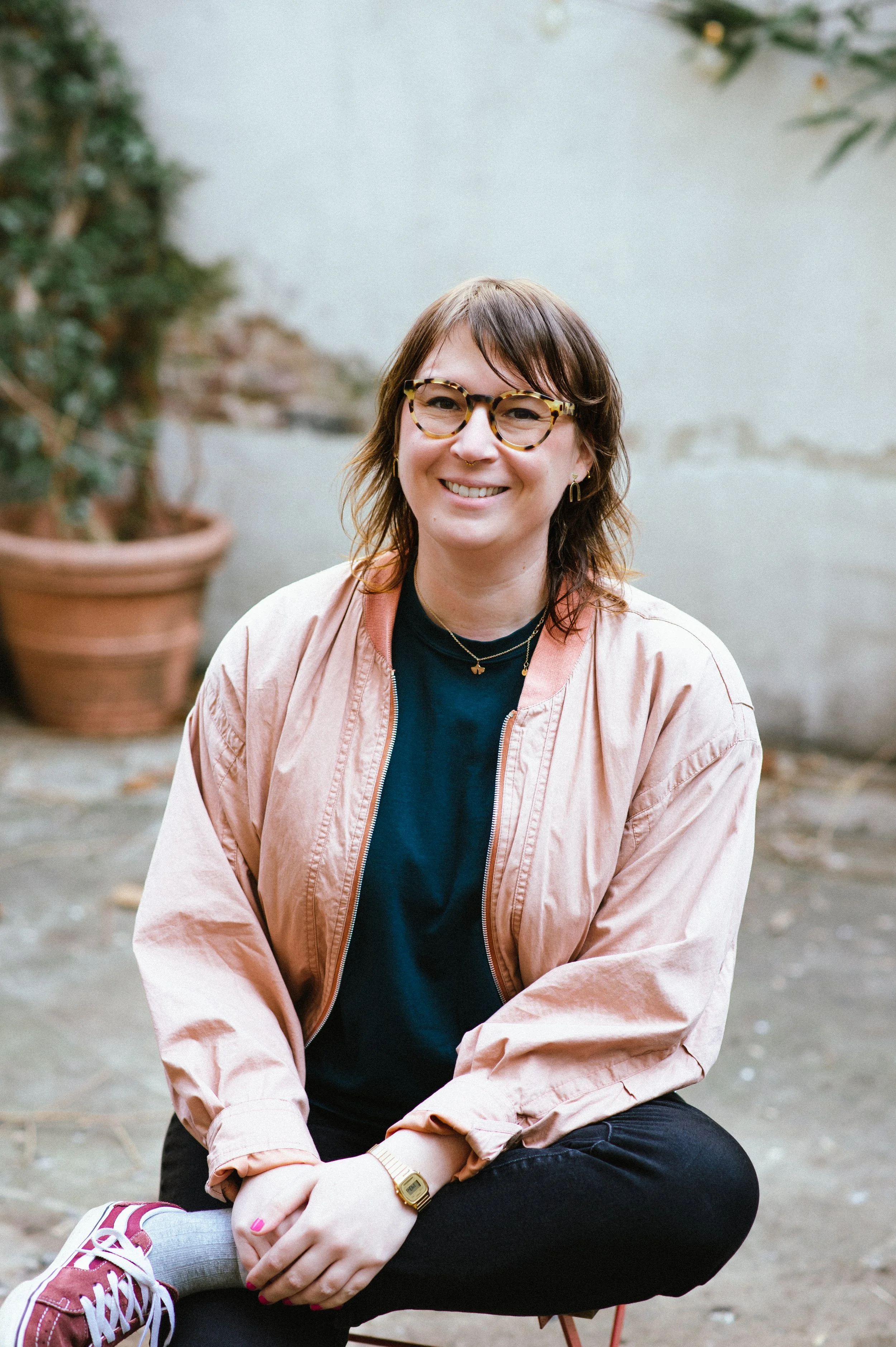 Junge Frau mit Brille sitzt draußen vor einer Wand, trägt eine rosa Jacke, ein schwarzes Oberteil, schwarze Hosen und Sneakers, und trägt Schmuck sowie eine Armbanduhr.