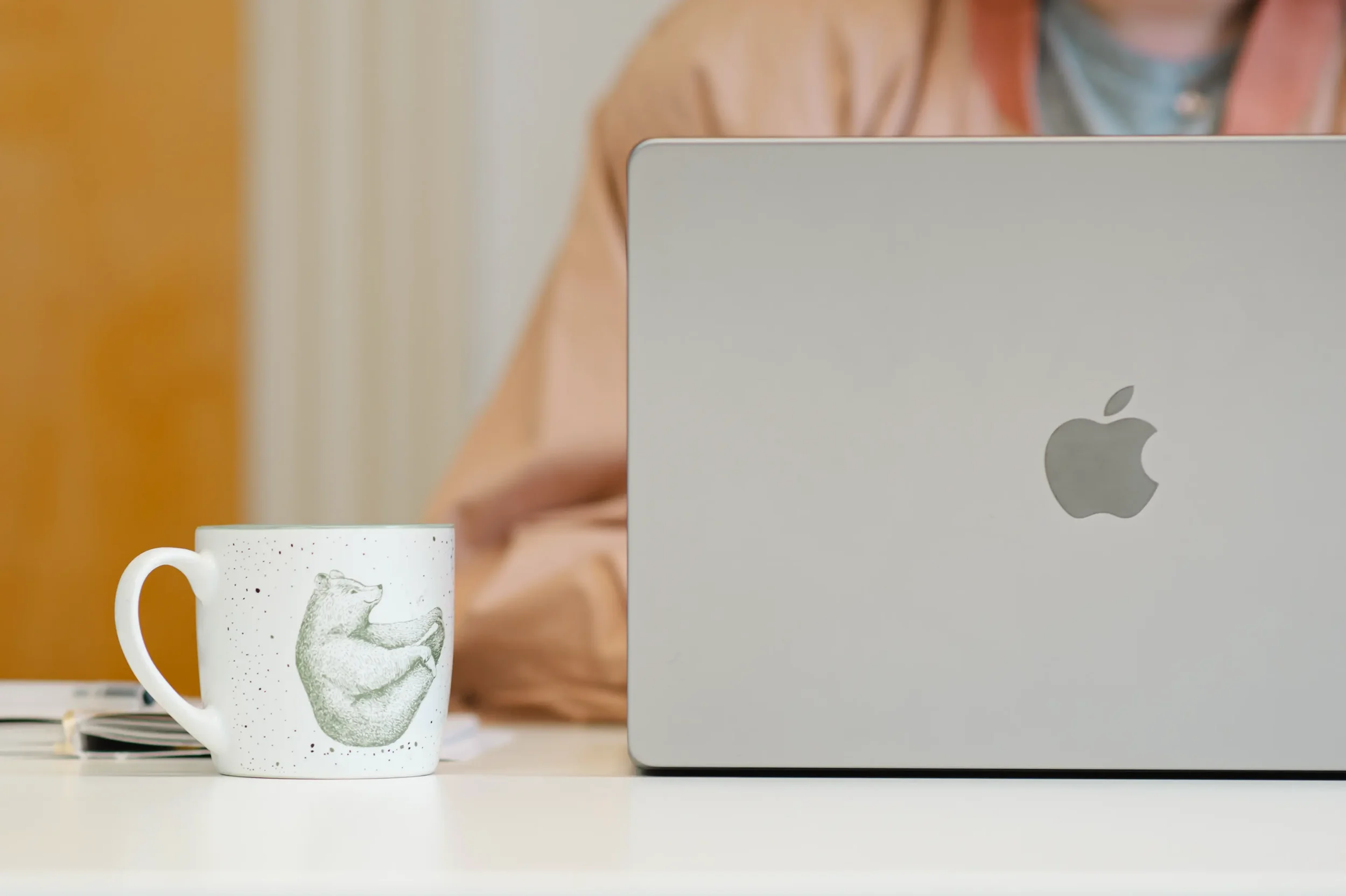 Eine weiße Kaffeetasse mit einem Bären, der einen Bananenstiel hält, auf einem weißen Tisch. Im Hintergrund ein Laptop und eine Person, die auf das Gerät schaut.