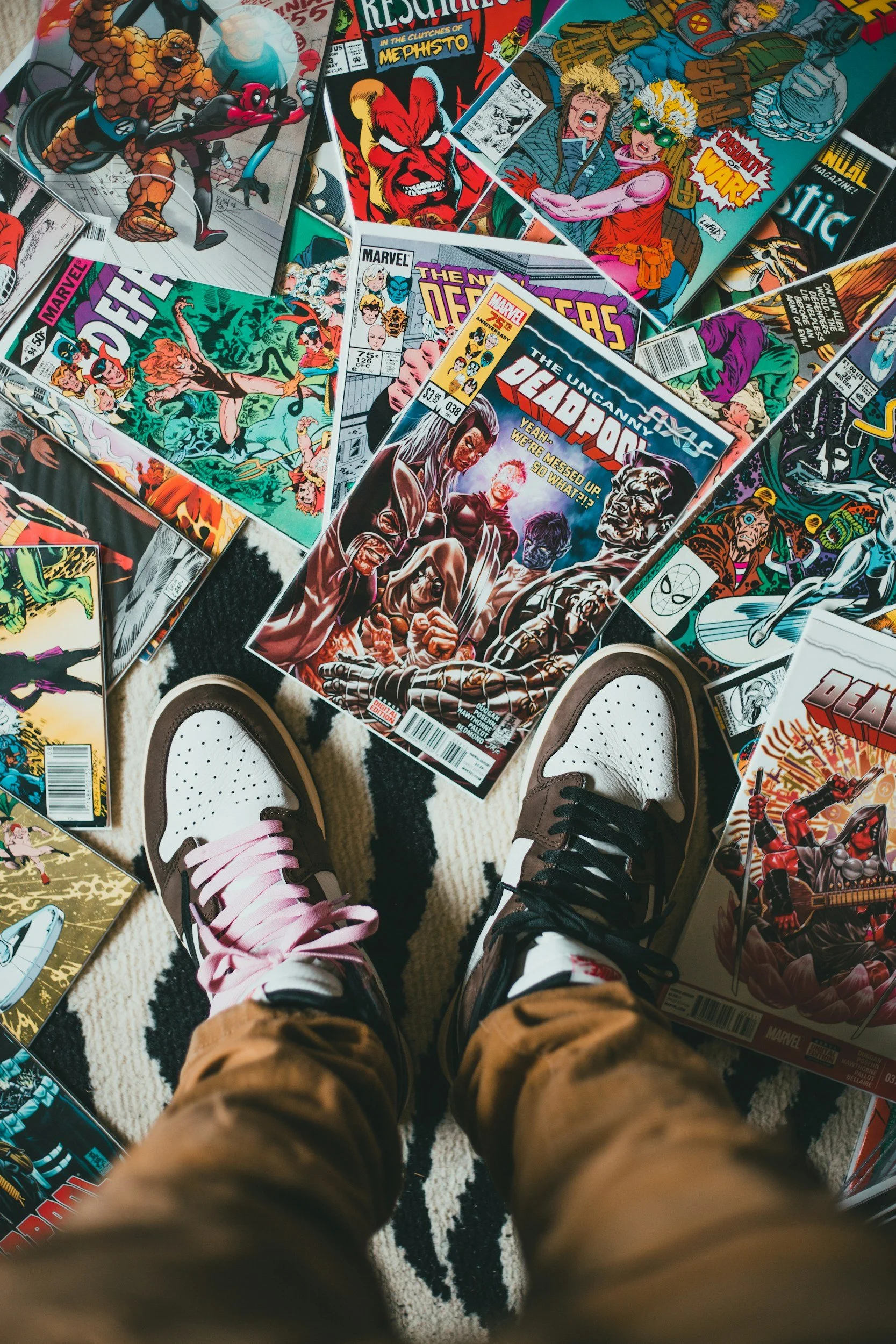 View of a person's legs and feet standing on a black and white patterned rug, surrounded by various Marvel comic books on the floor, including Deadpool, The X-Men, and The Avengers, with the person wearing sneakers, one pink and white and the other black and white.