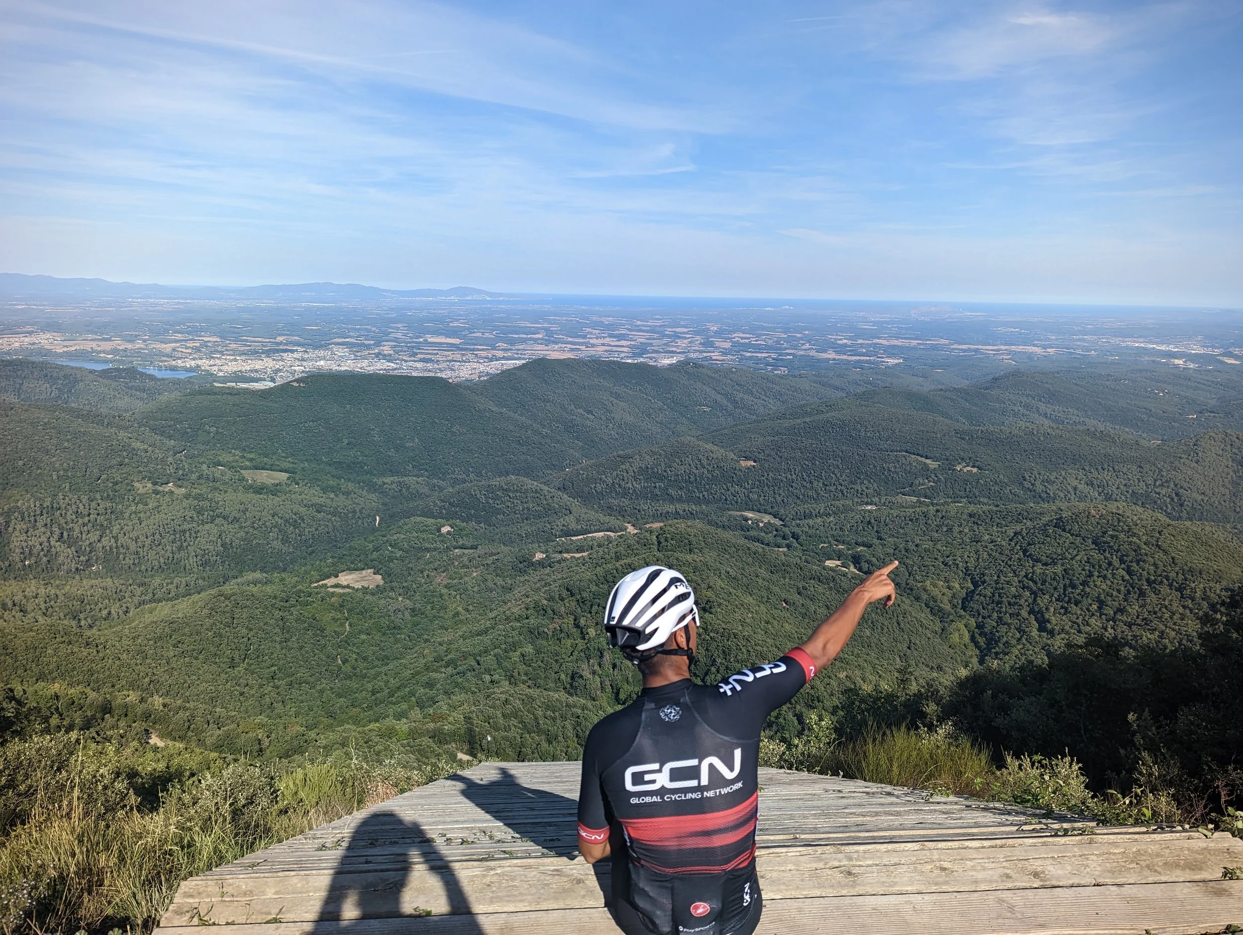 A cyclist wearing a black and red jersey with 'GCN' on the back pointing towards the horizon as they stand on a wooden trail, with green hills and a vast landscape in the background under a partly cloudy sky.