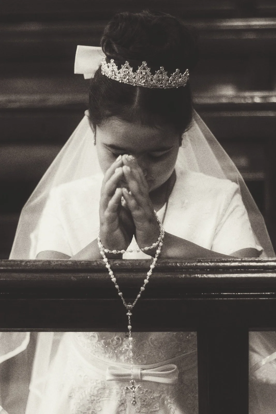 A woman kneeling in prayer with her eyes closed, hands clasped, wearing a crown, veil, and rosary in a church or chapel.