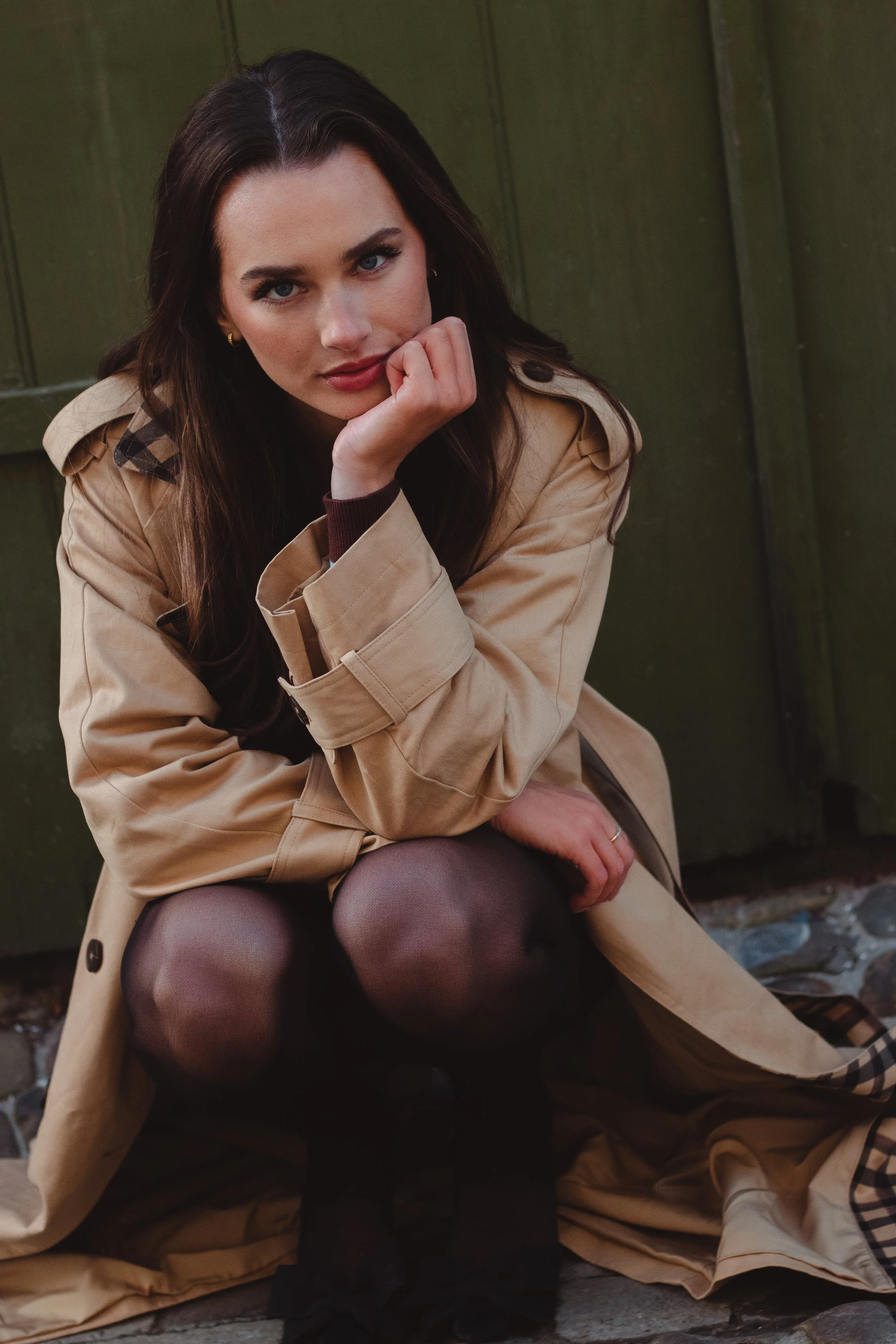 A woman with dark brown hair and blue eyes sitting outdoors against a green wooden wall, wearing a tan trench coat, black tights, and dark shoes, with her chin resting on her hand and a contemplative expression.