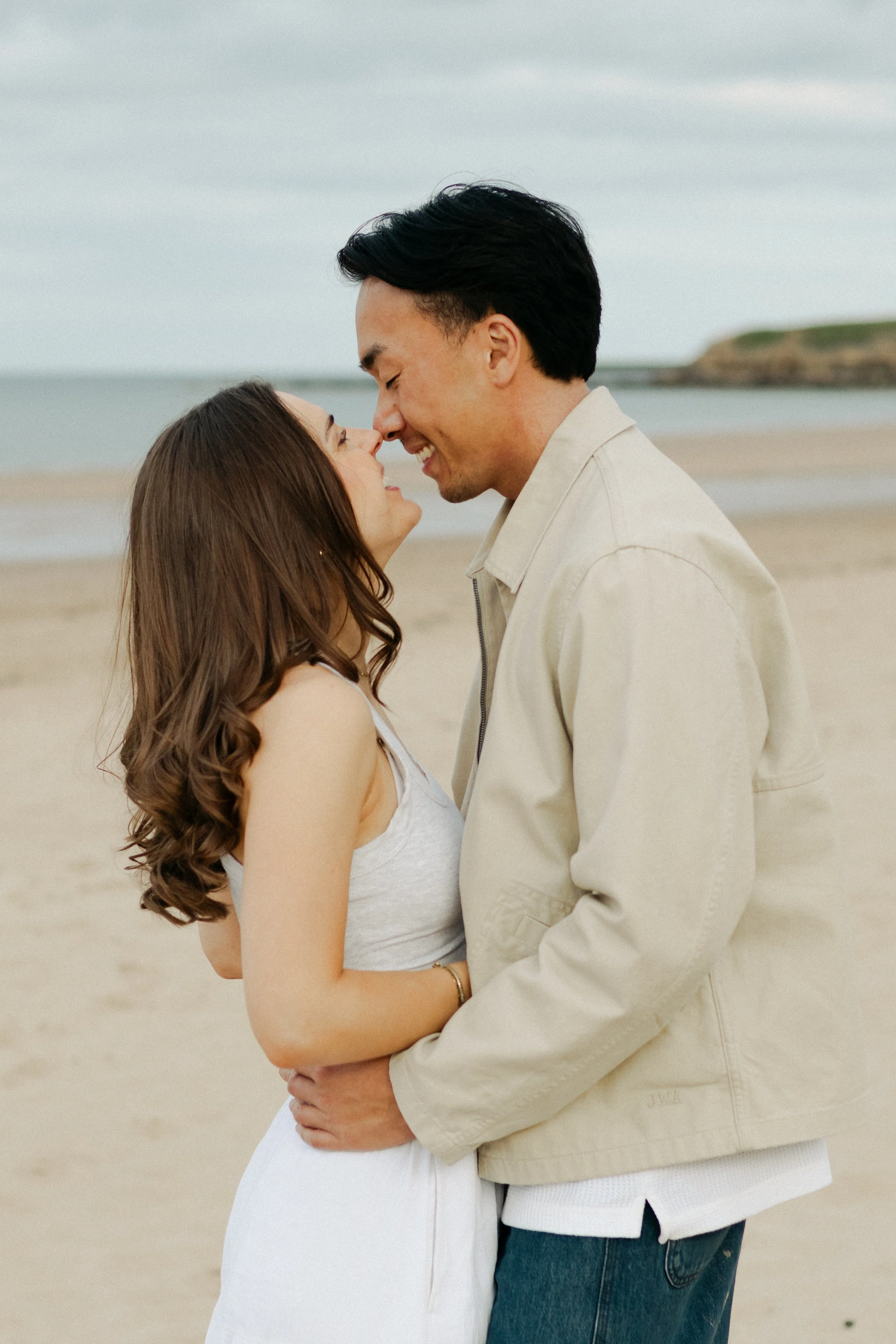 A couple stands close together on a beach, touching foreheads and smiling at each other.