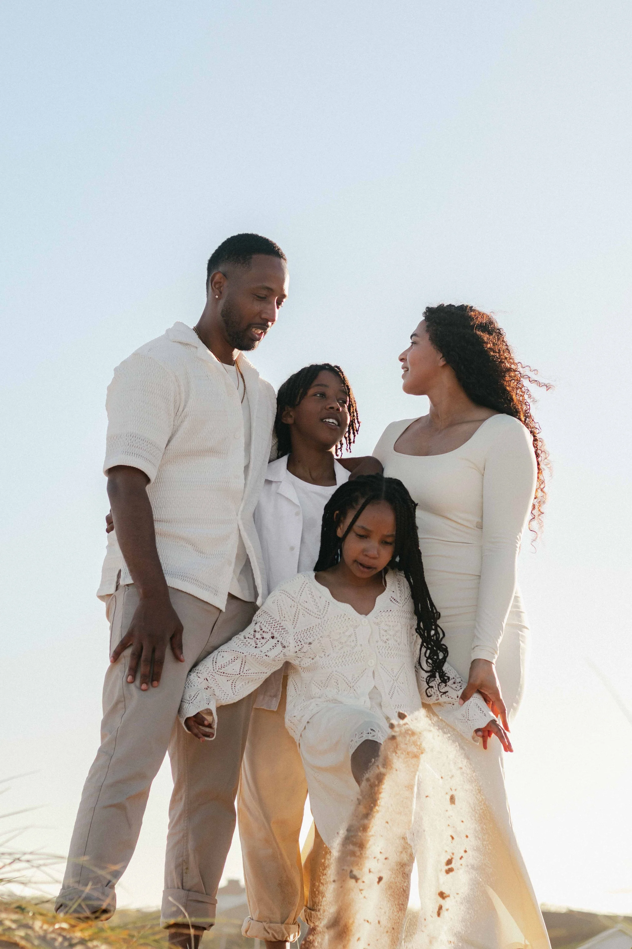 Group of African American family members enjoying together outdoors, with a woman in a white dress and children around her, during sunset or late afternoon.