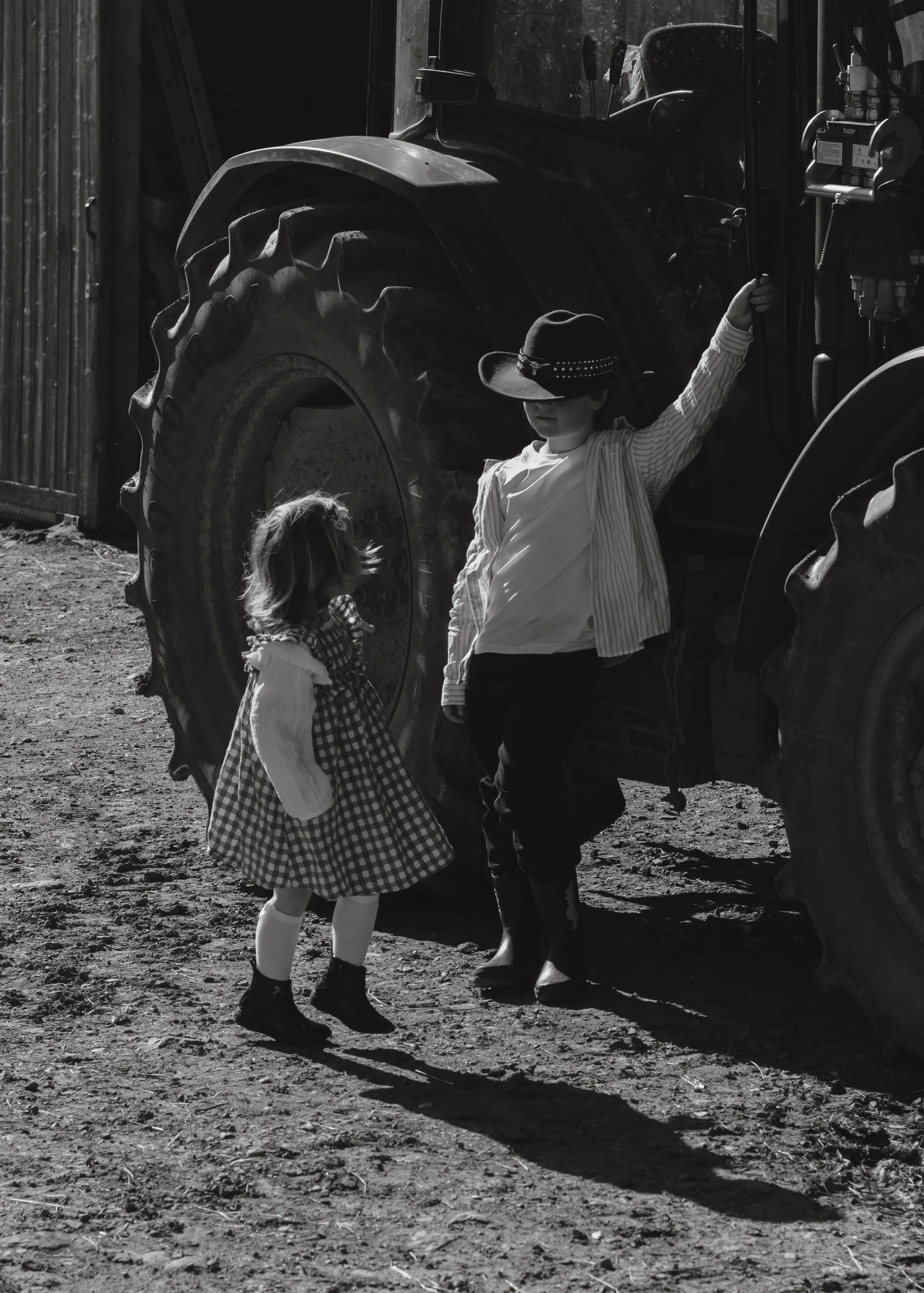 Two children, a boy and a girl, standing near a large tractor. The boy is wearing a cowboy hat and a striped shirt, holding onto part of the tractor. The girl is wearing a checkered dress, looking up at the boy. The scene is outdoors with dirt ground, and the image is in black and white.