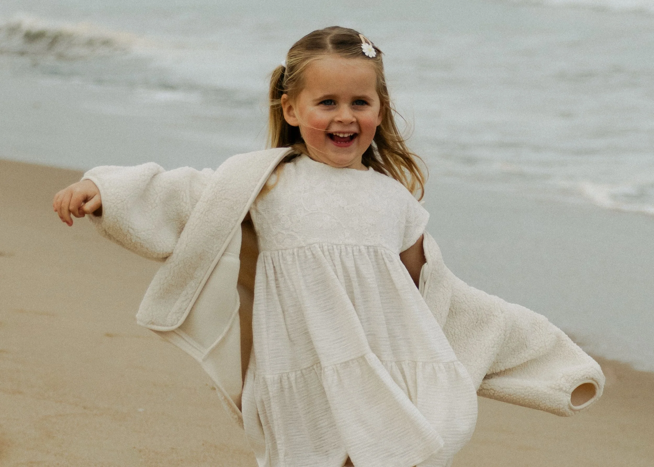 A young girl with long blonde hair, wearing a cream-colored dress and a matching jacket, standing on a beach with the ocean in the background, smiling and looking happy.