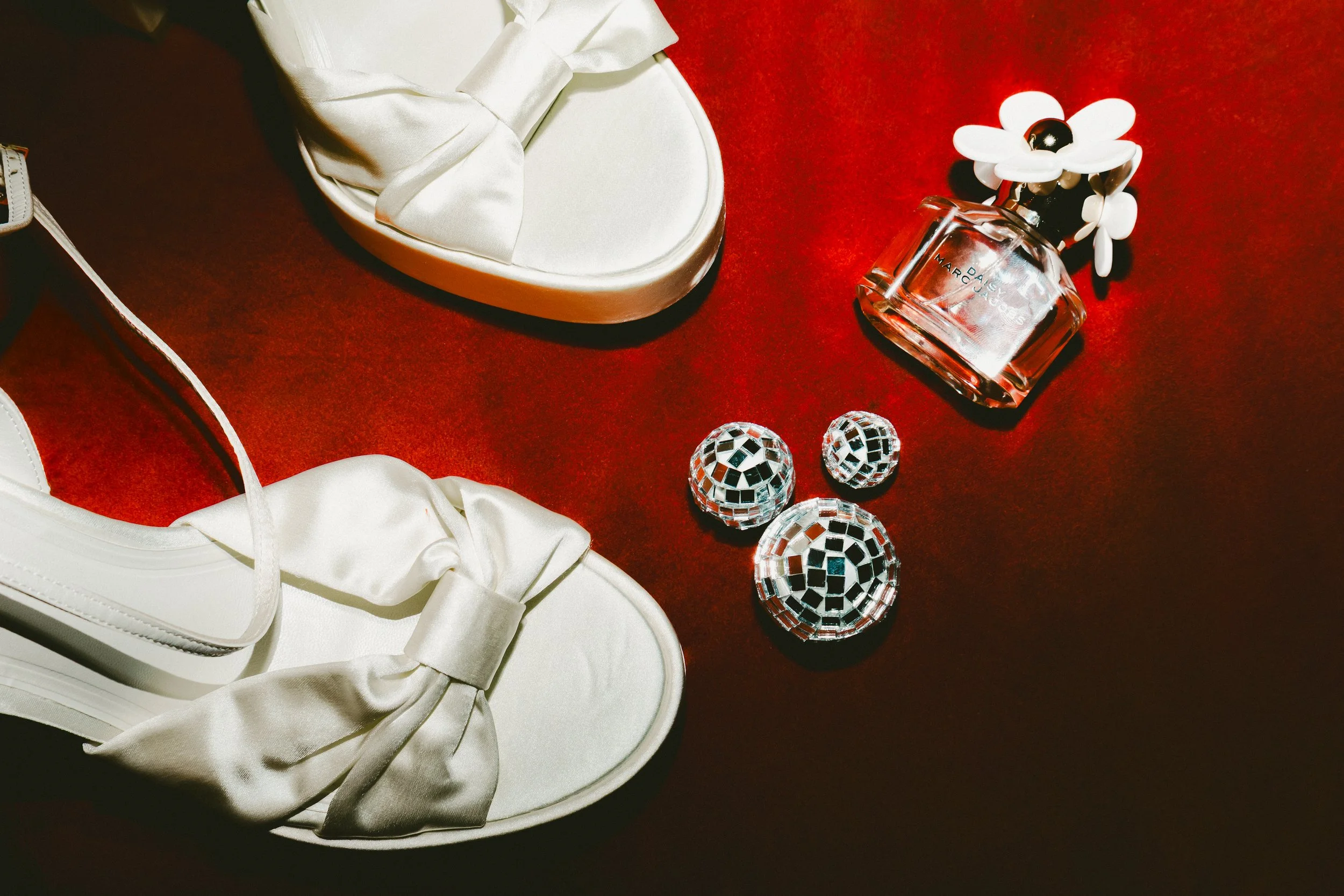 A white satin shoe with a bow on a red surface, positioned near a white box with a satin lining. To the right, there is a glass perfume bottle decorated with a white flower cap, and three small mirrored decorative spheres.