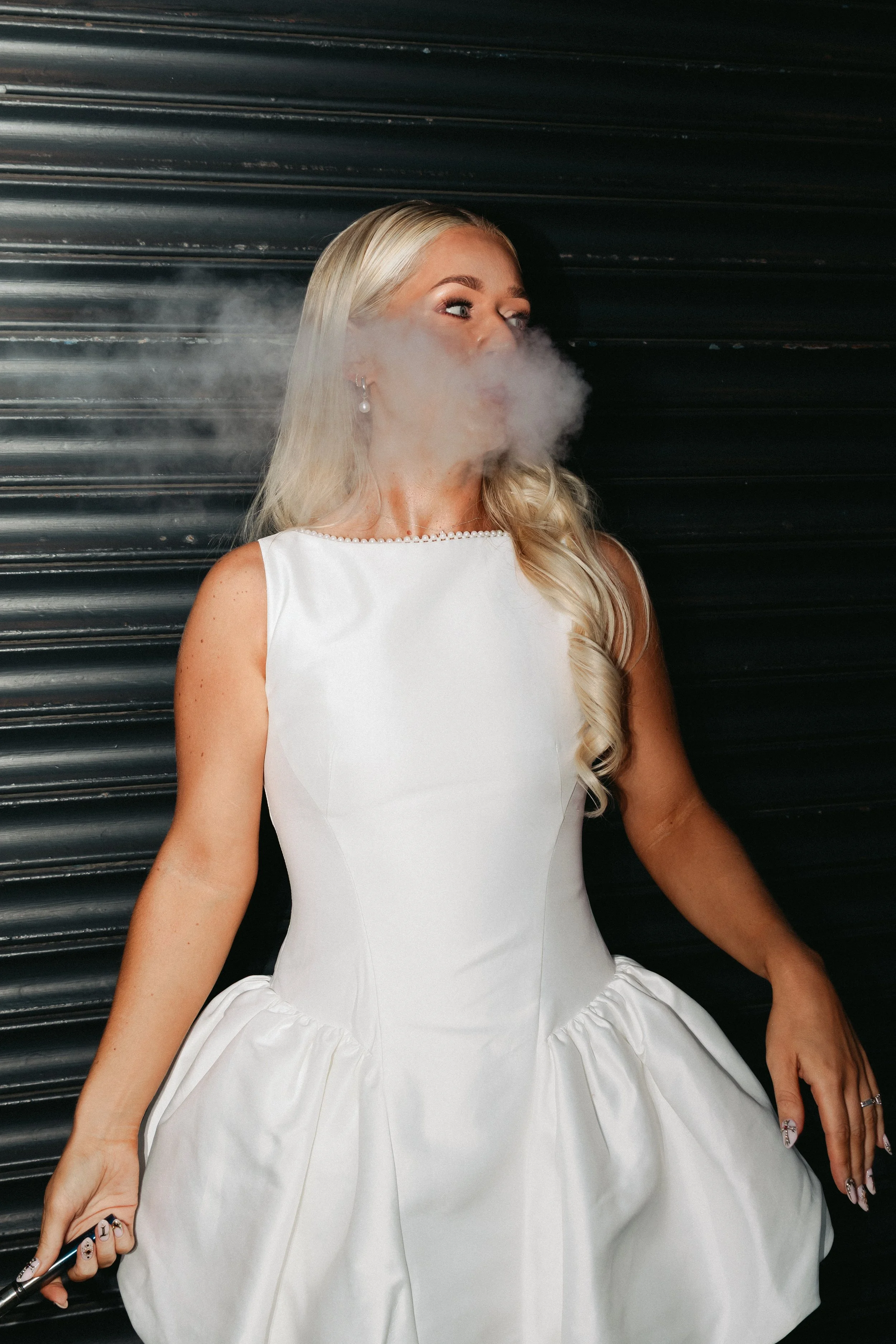 A blonde woman in a white dress with pearl earrings and rings, smoking and sitting against a black, ridged background.