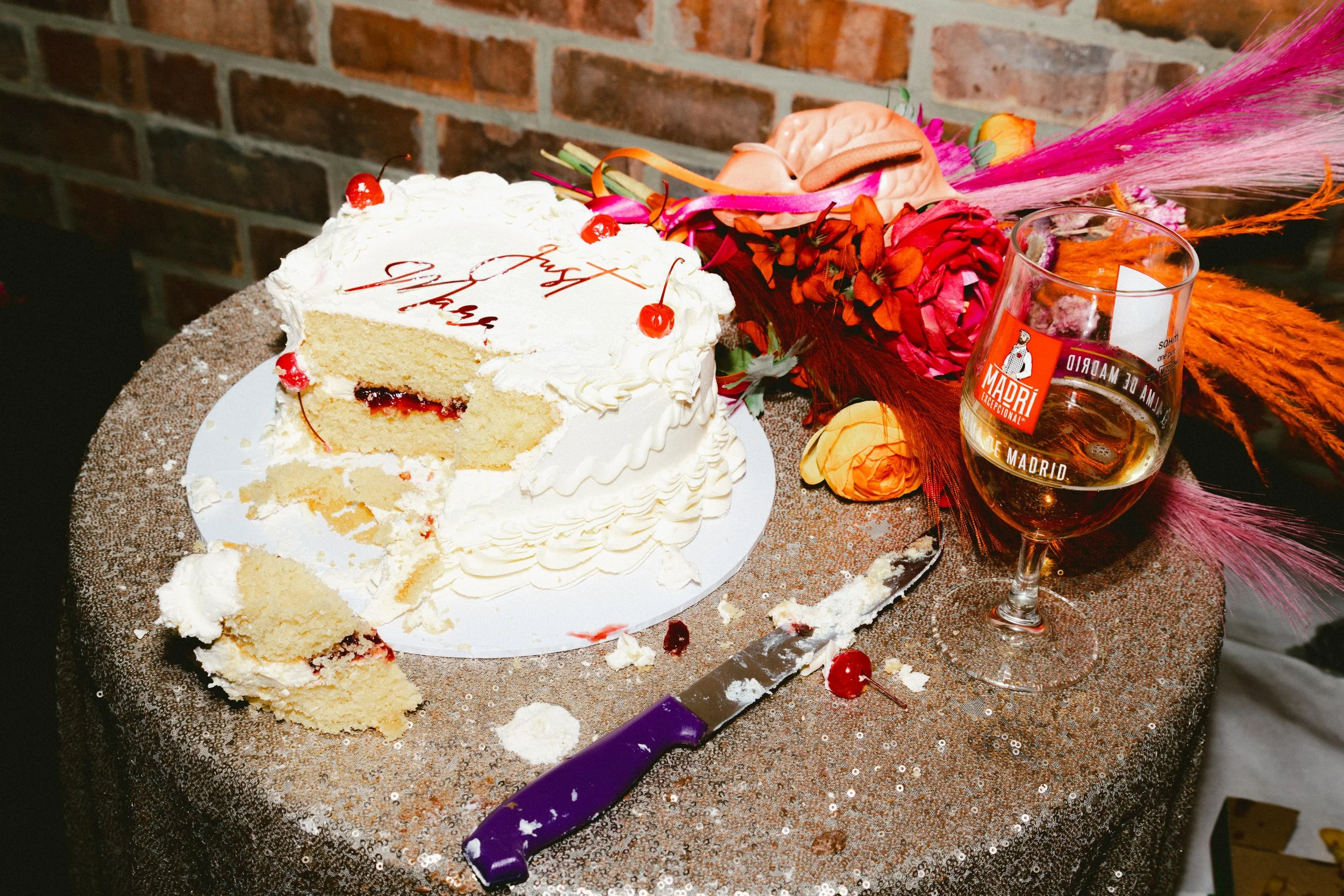 A partially eaten white birthday cake with cherries on top, a glass of amber-colored beverage with a red logo, and a floral arrangement on a glittery gold table against a brick wall background.