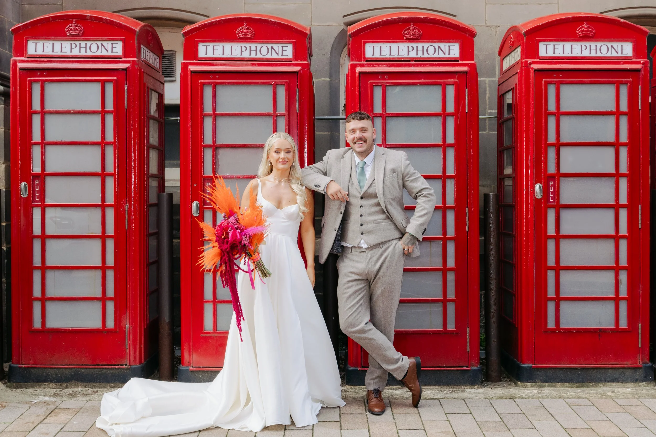 A bride and groom posing in front of four red British telephone booths. The bride is holding a bouquet of bright pink and orange flowers and is wearing a white wedding dress. The groom is dressed in a light gray suit with a mint green tie, smiling and leaning on the phone booth.