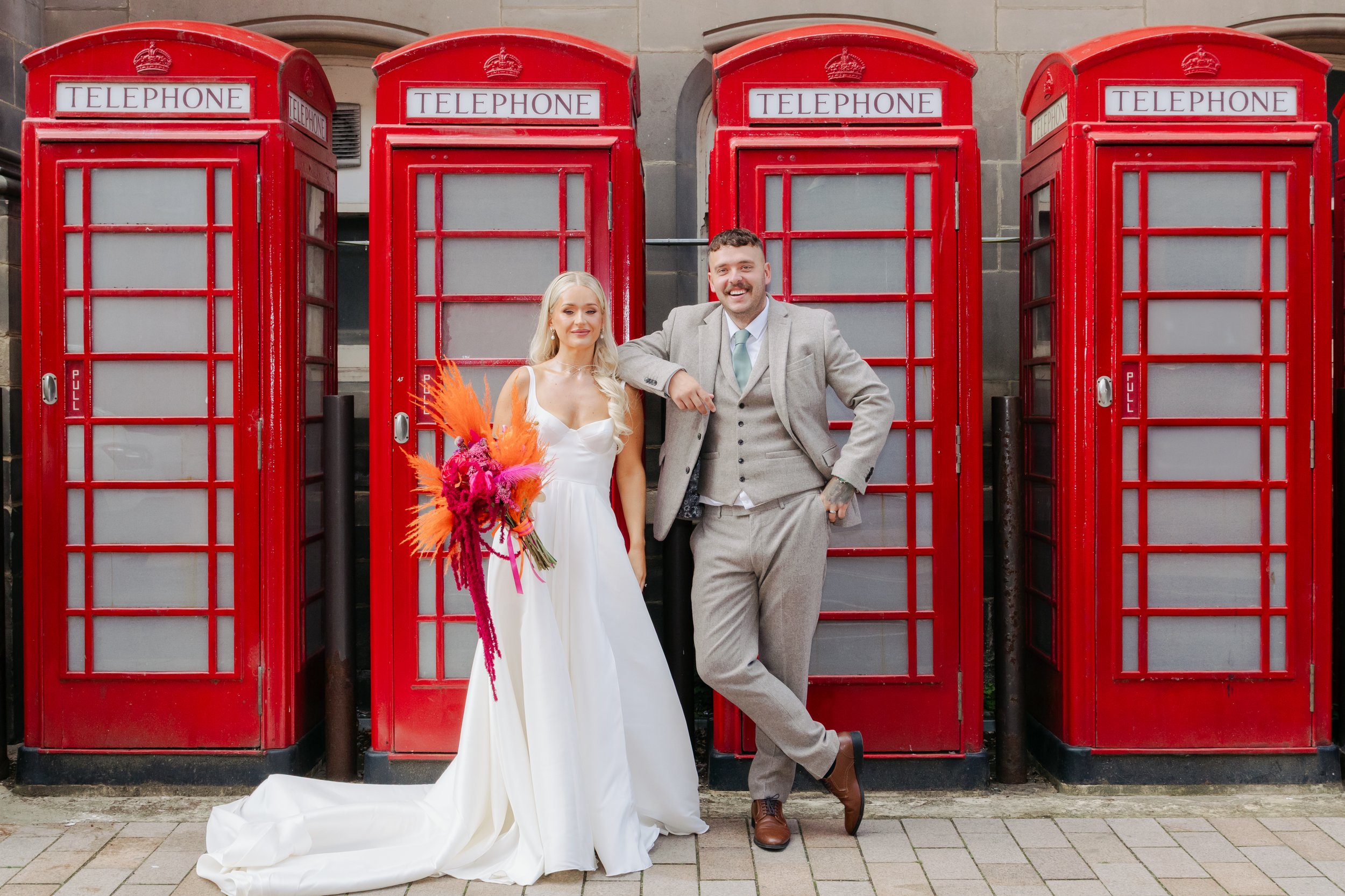 A bride and groom posing in front of four red British telephone booths. The bride is holding a bouquet of bright pink and orange flowers and is wearing a white wedding dress. The groom is dressed in a light gray suit with a mint green tie, smiling and leaning on the phone booth.