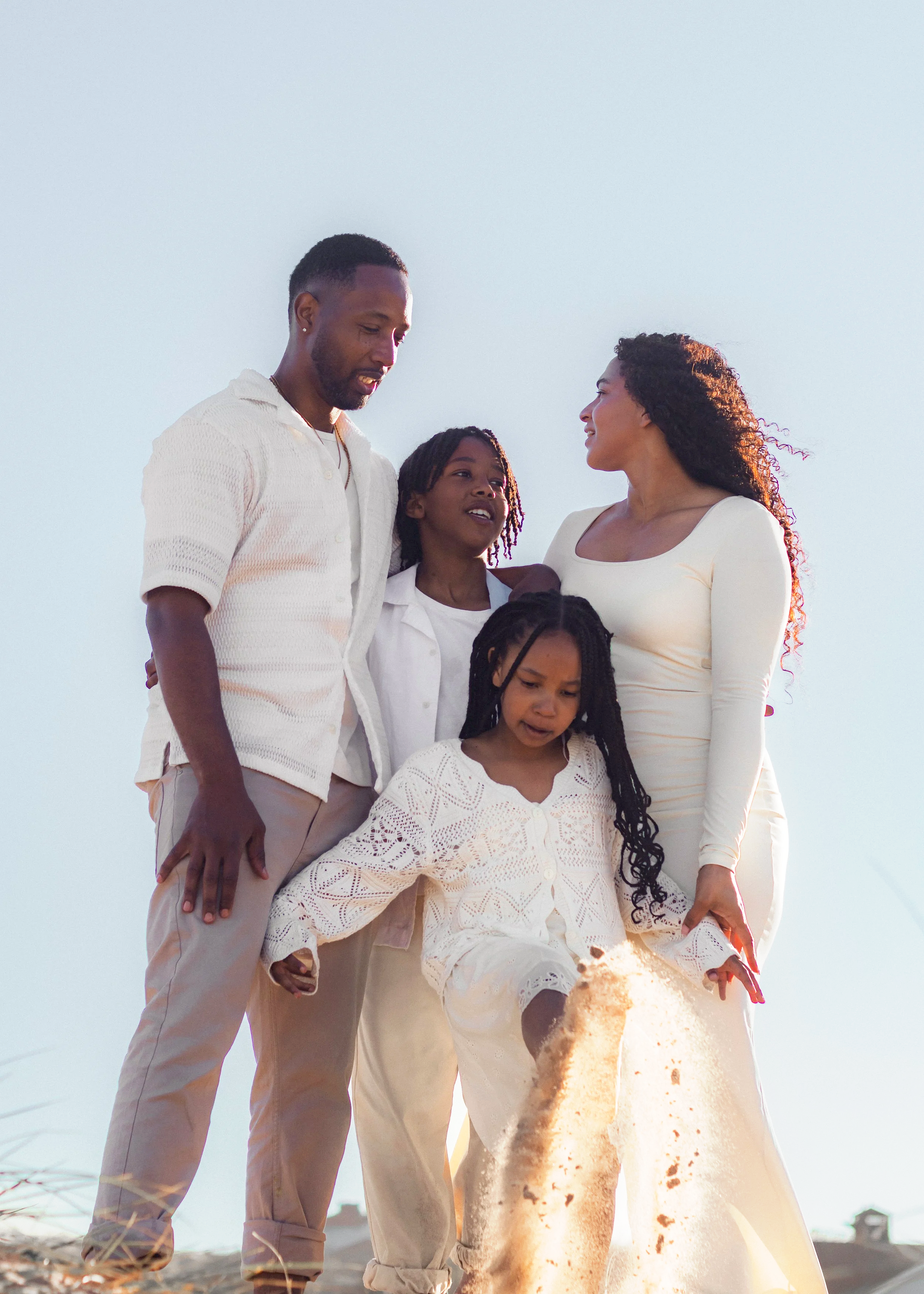 A family of four celebrating outdoors during a sunny day on a beach, with the little girl kicking sand