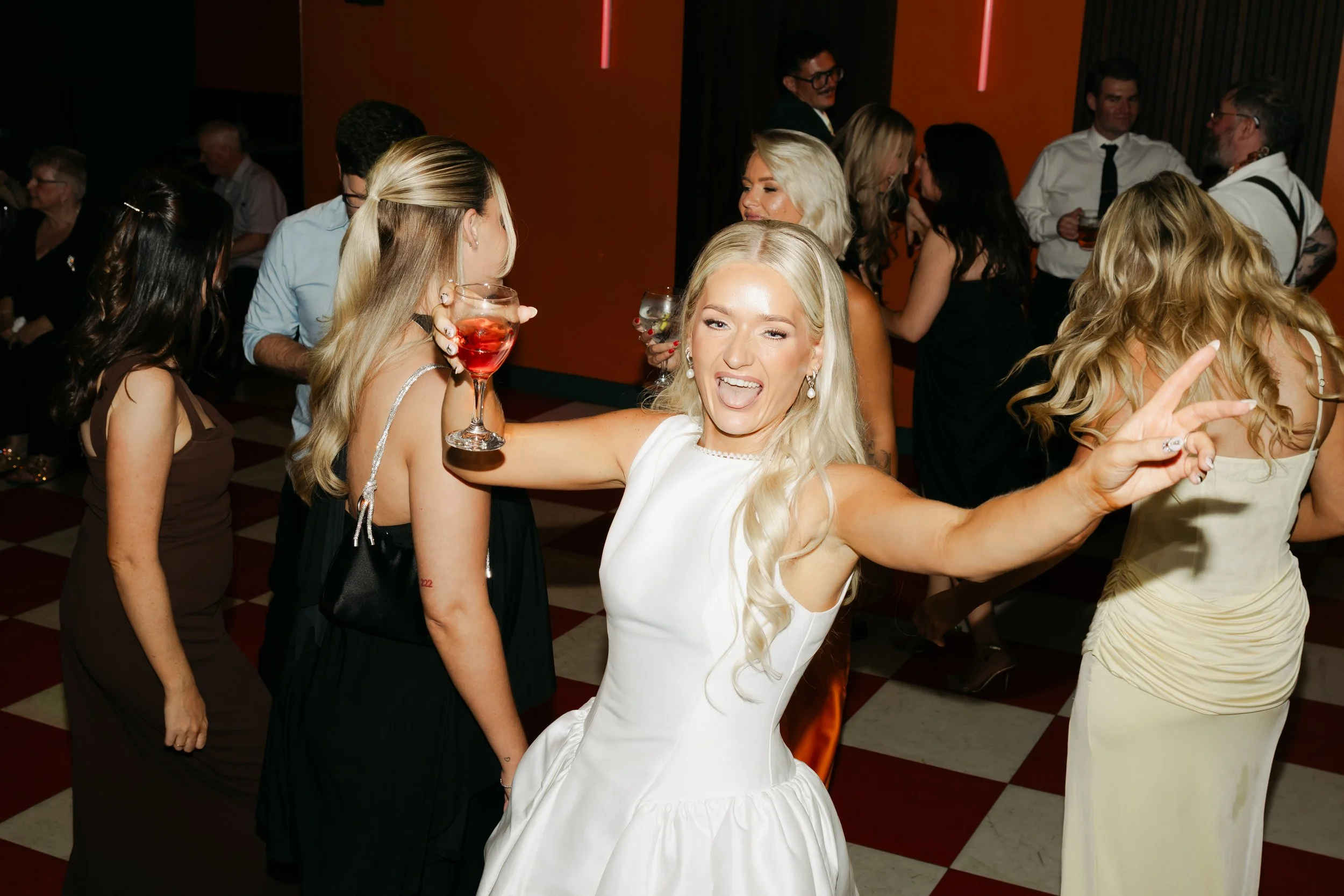 A group of people dancing and socializing at a party. A blonde woman in a white dress is smiling and making a peace sign.