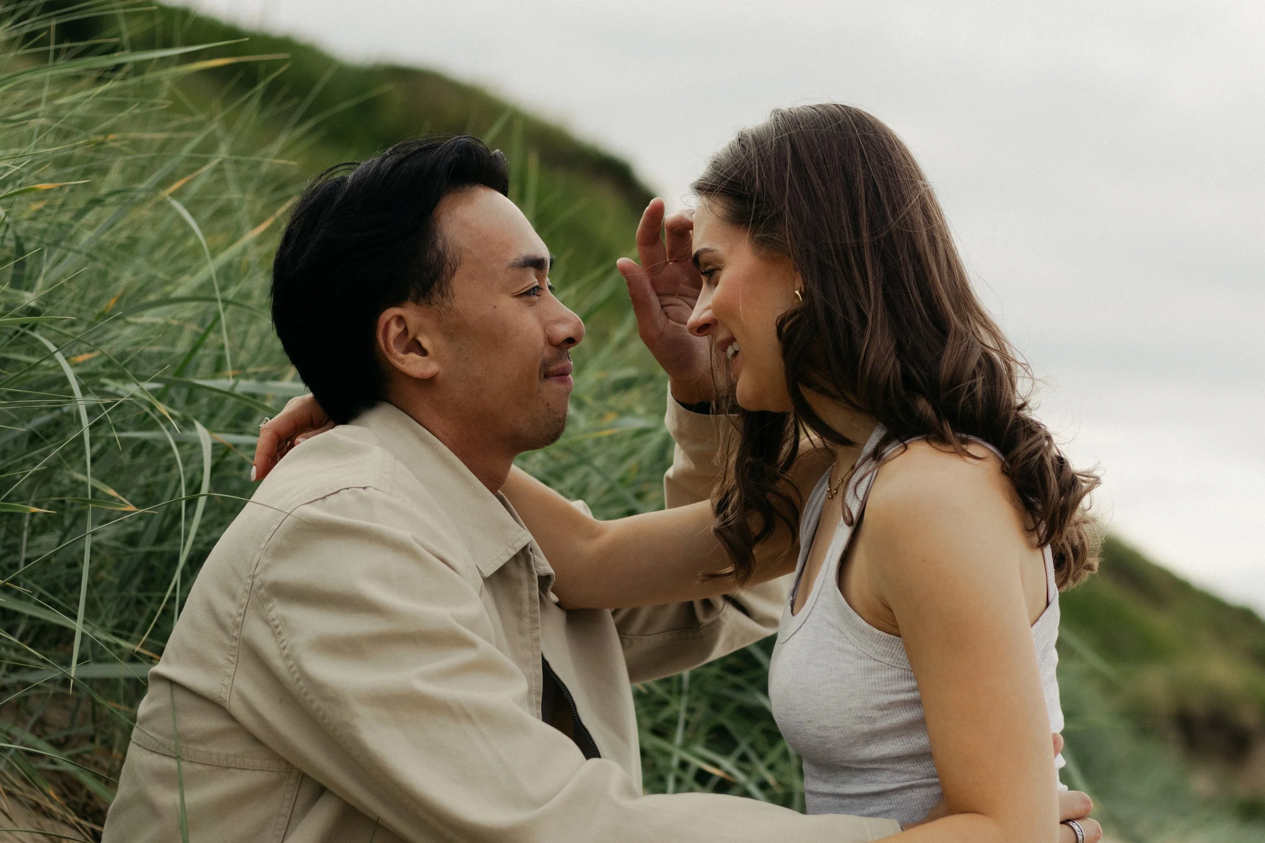 A man and a woman are sitting close together outdoors amidst tall green grass, facing each other with affectionate expressions.