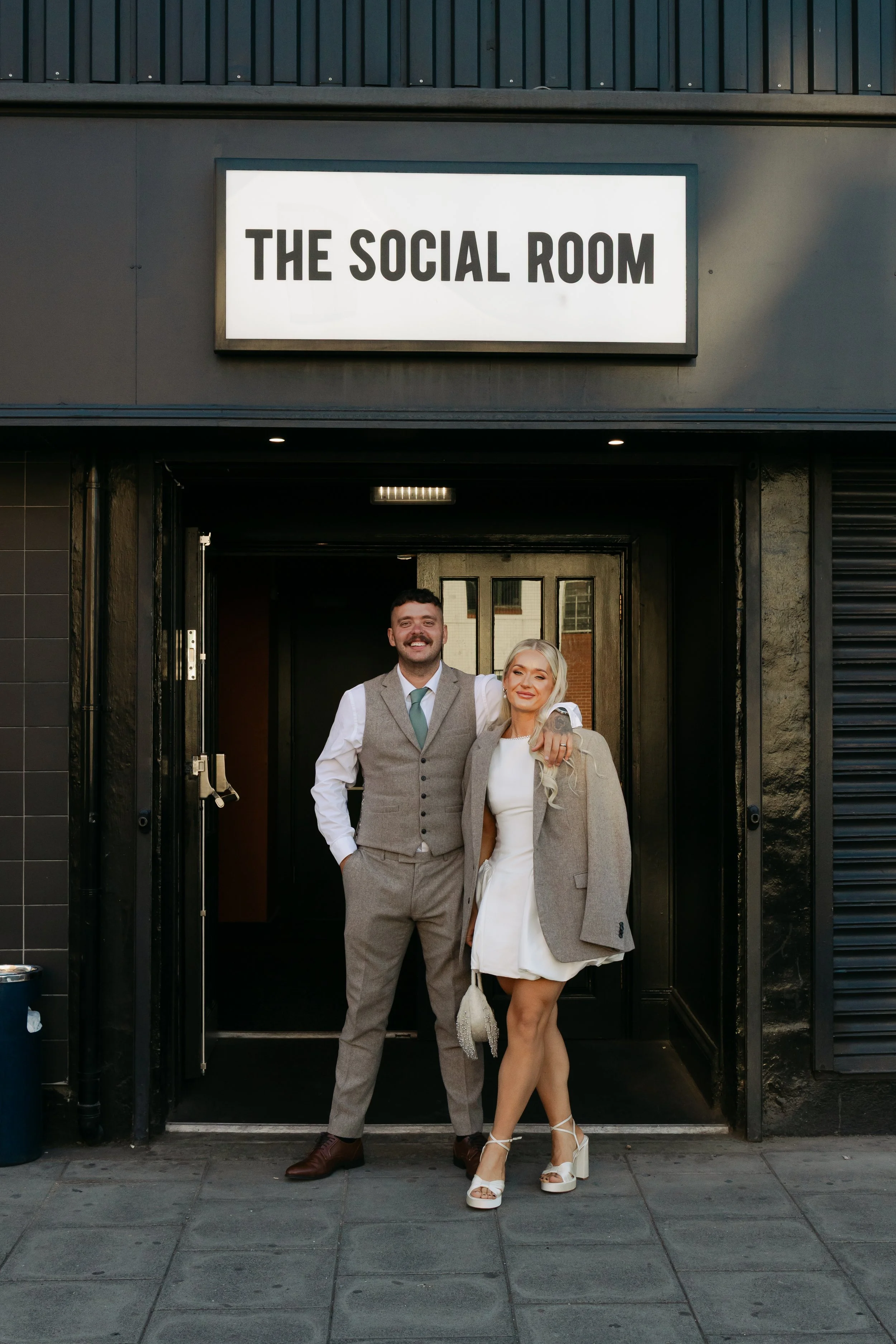 Two people, a man in a gray suit and a woman in a white dress with high heels, standing arm in arm in front of an entrance with a sign that says "The Social Room."