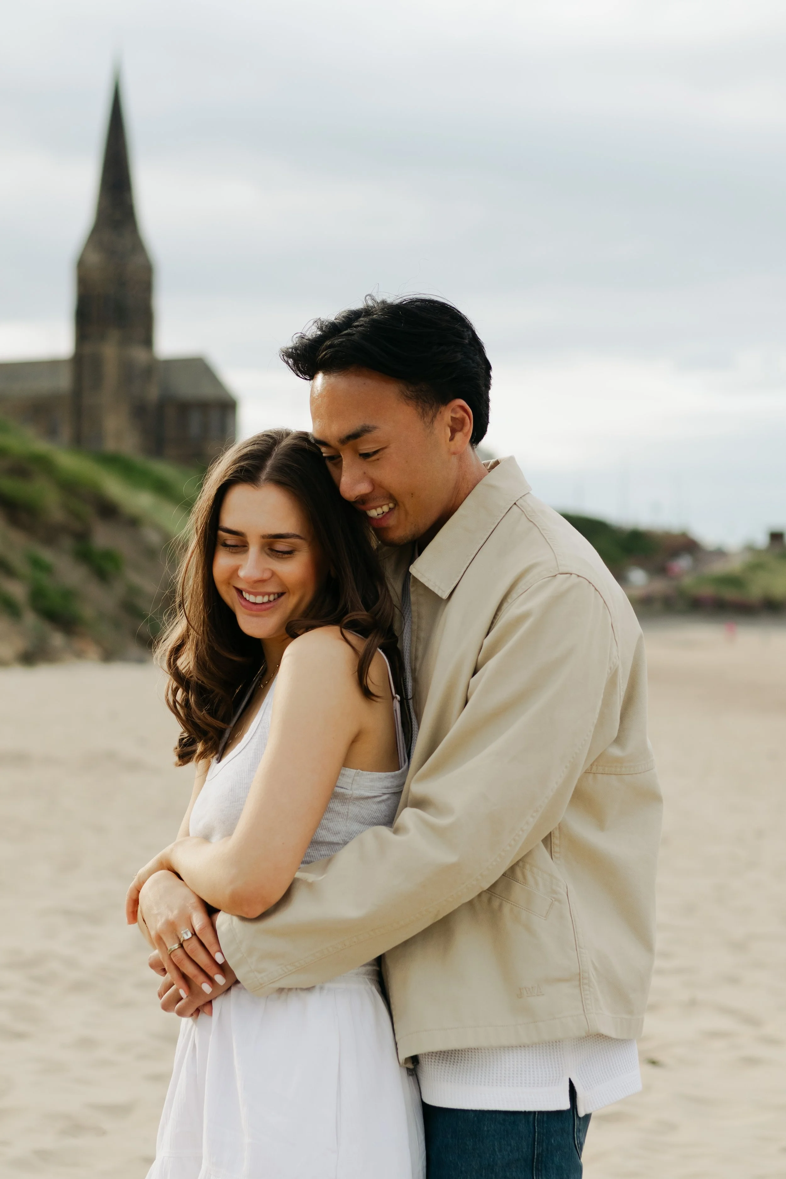A couple embracing on a beach with a church in the background, smiling and enjoying each other's company.