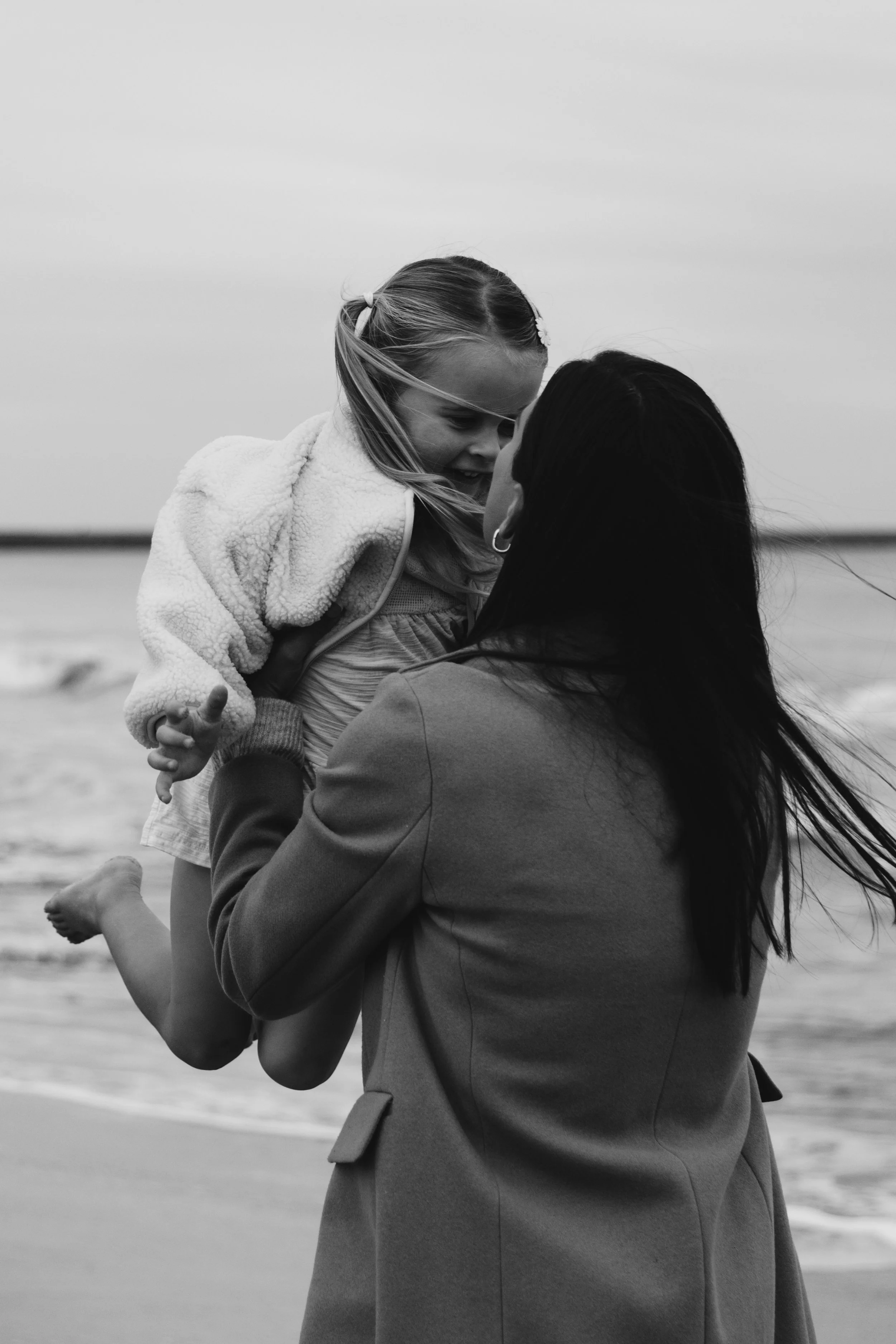 A woman holding a smiling young girl with long hair at the beach, both facing each other. The woman is wearing a blazer, and the girl has a hooded jacket. The background shows a beach with the ocean.