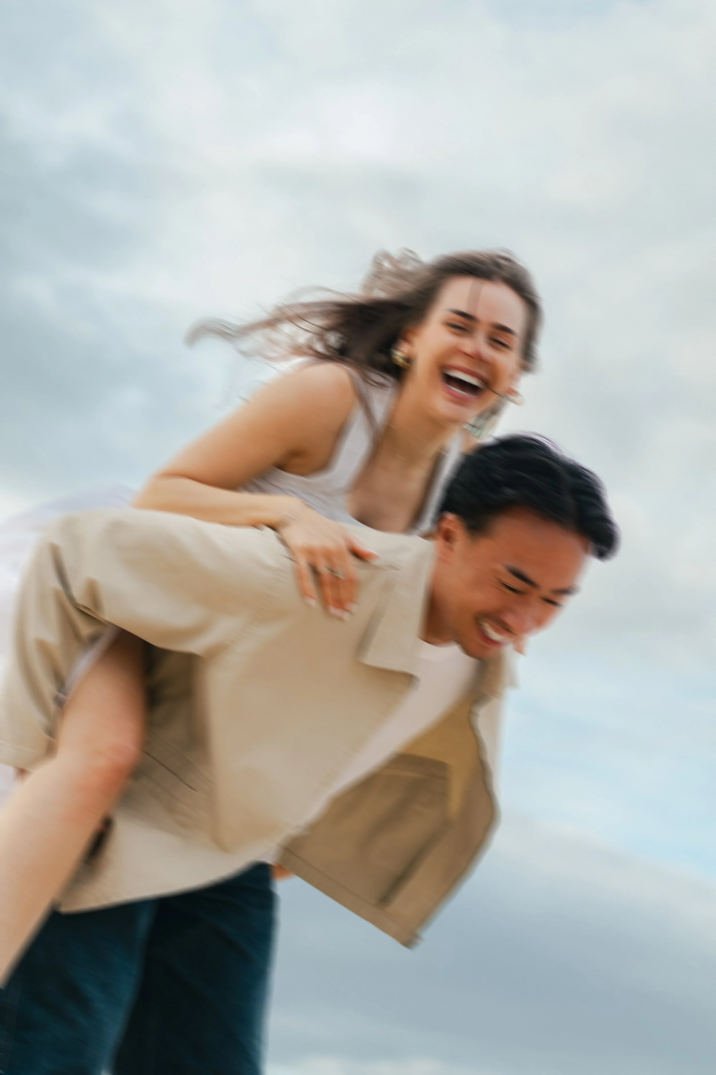 A woman with long brown hair laughing while riding on a man's back, who is carrying a box, outdoors against a cloudy sky.