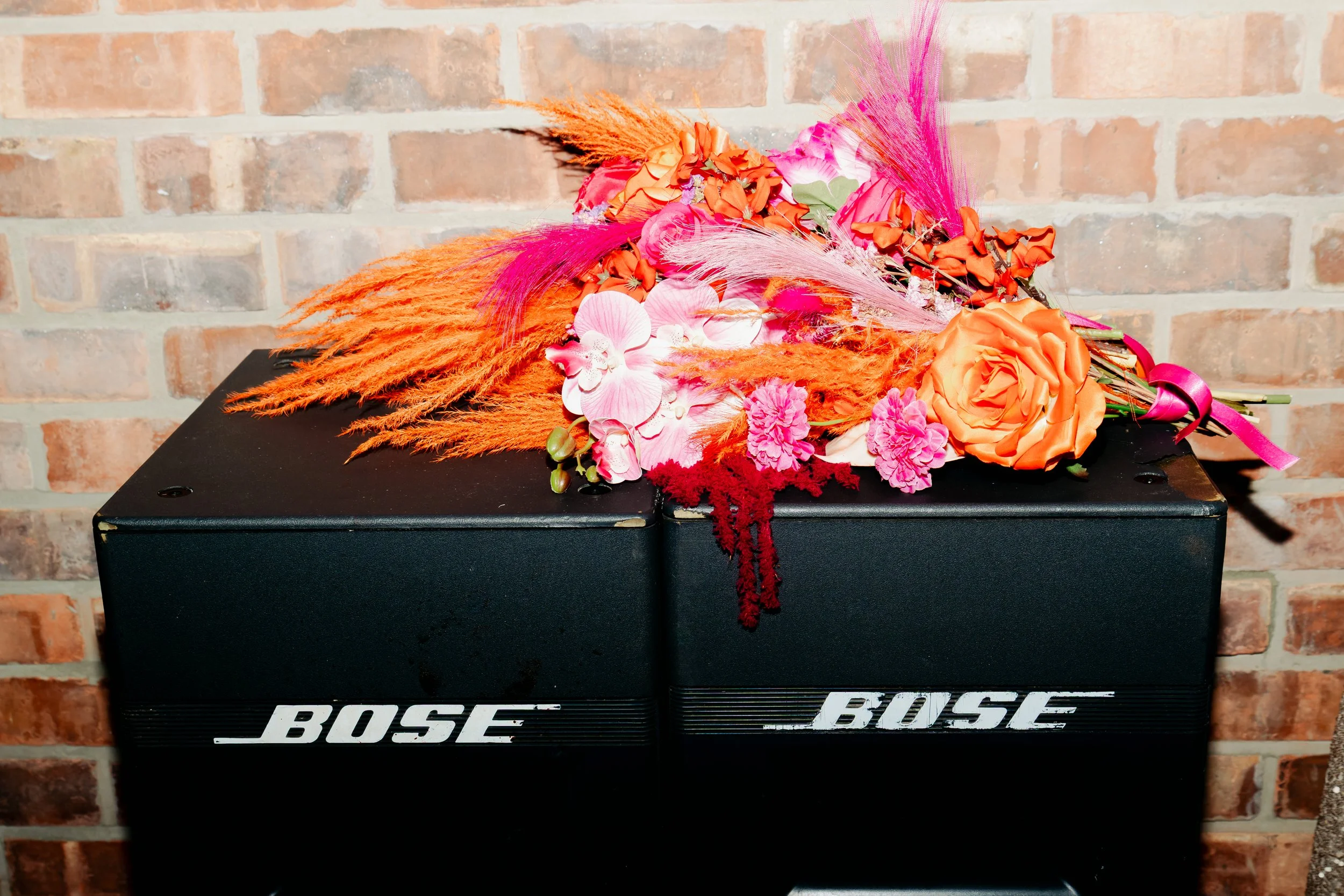 A colorful flower arrangement with pink, orange, and red flowers and feathers resting on top of a black Bose speaker.