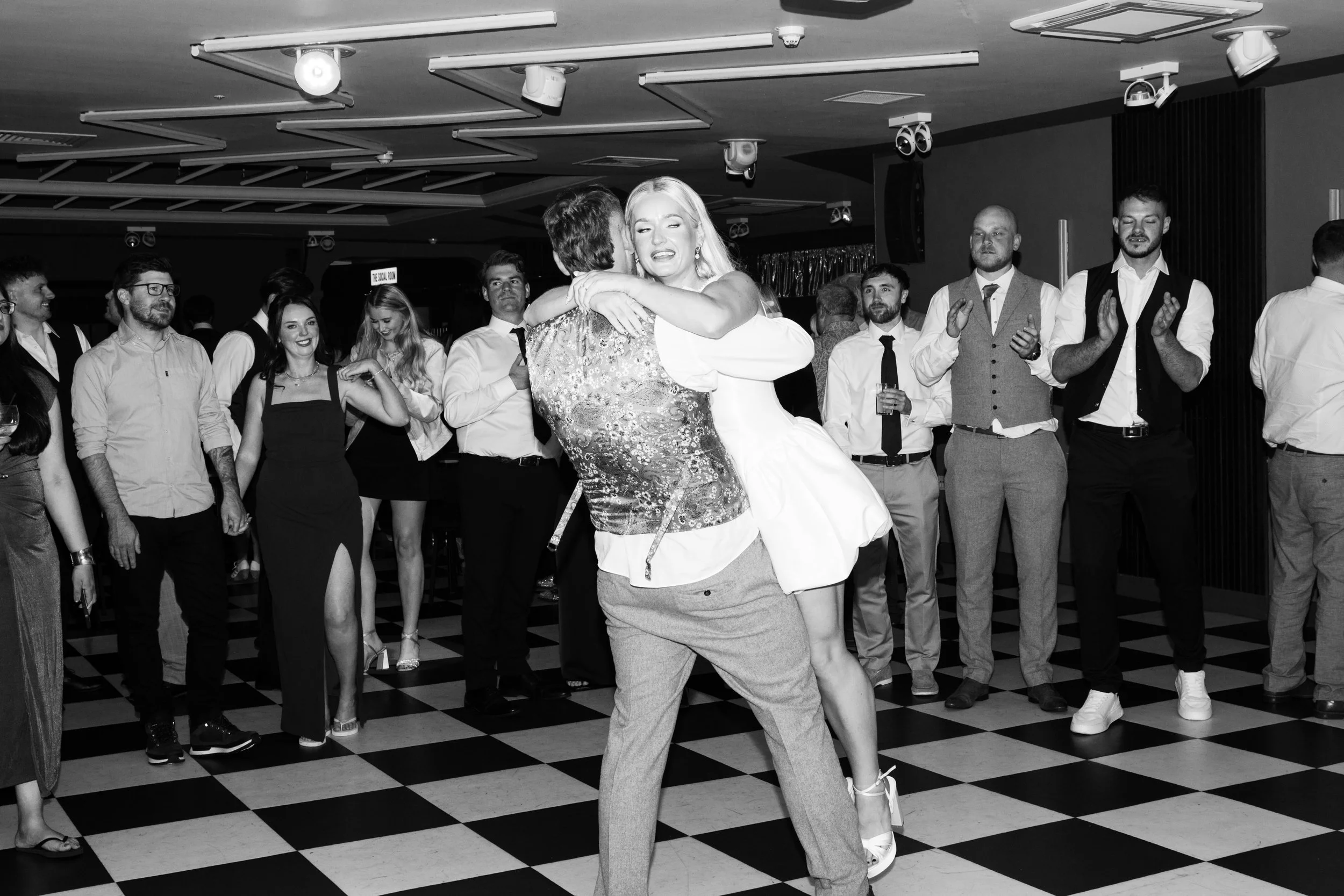 A woman in a white dress and a woman in patterned clothing dance closely at a party with people watching and clapping on a checkered floor.