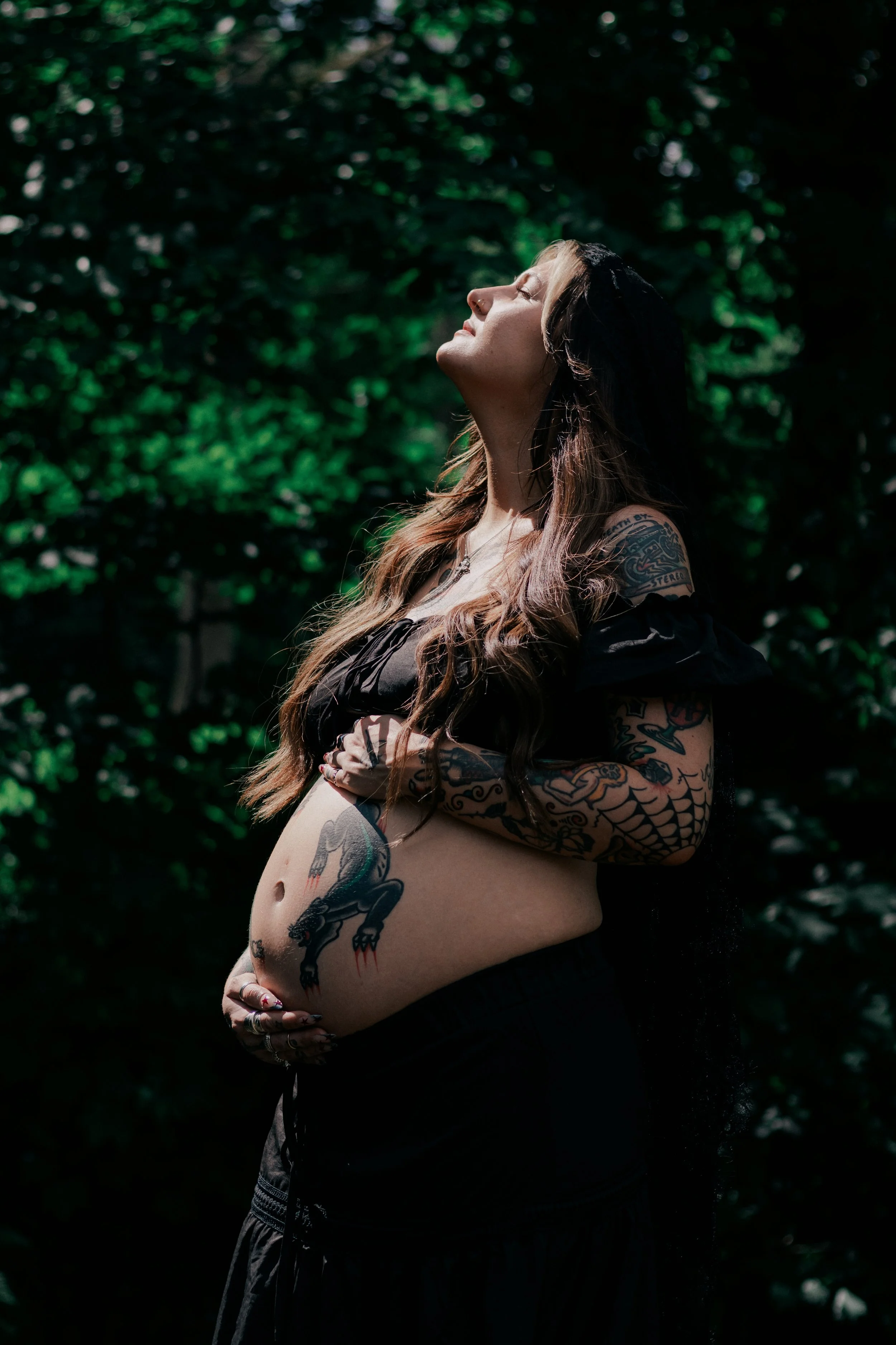 A pregnant woman with tattoos on her arms and stomach, standing with her eyes closed and face tilted upward, in a forested area with sunlight filtering through the greenery.