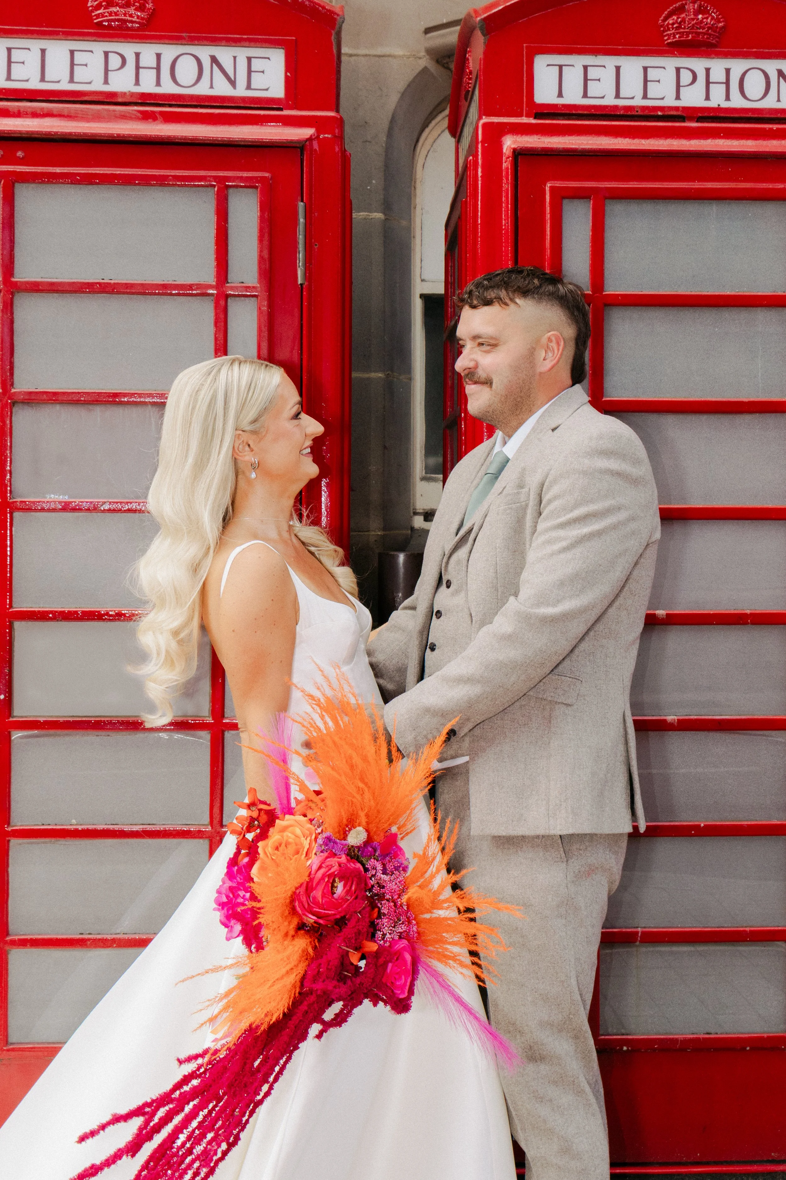 A bride and groom facing each other, holding hands, with a background of two red British telephone booths. The bride has long blonde hair and is wearing a white wedding dress, holding a colorful bouquet with orange and pink feathers and flowers. The groom is wearing a light gray suit and a pale blue tie.