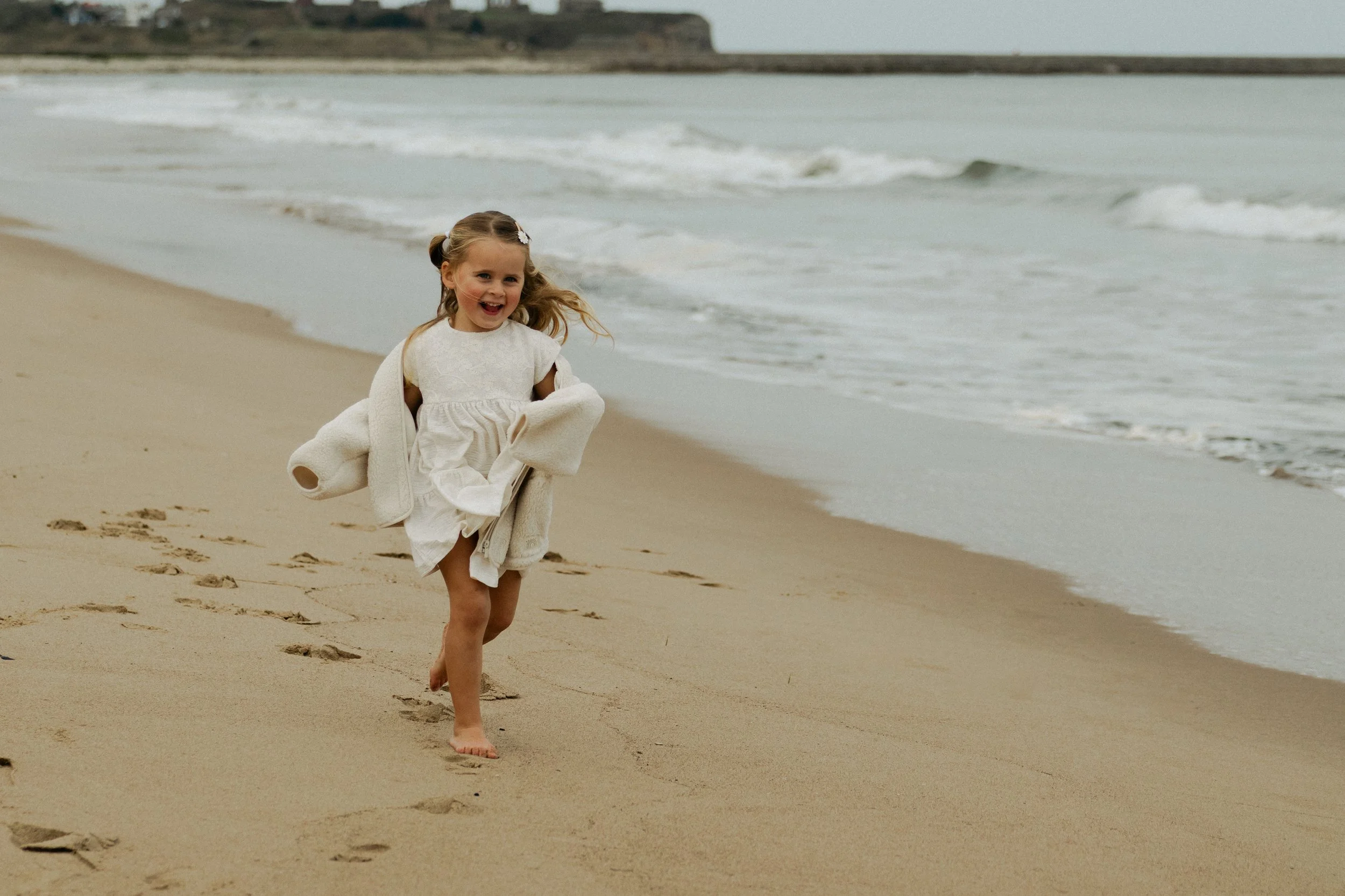 A young girl running barefoot on the sandy beach near the water's edge, smiling with her hair in pigtails, wearing a white dress and a beige jacket, with footprints in the sand behind her and waves in the background.