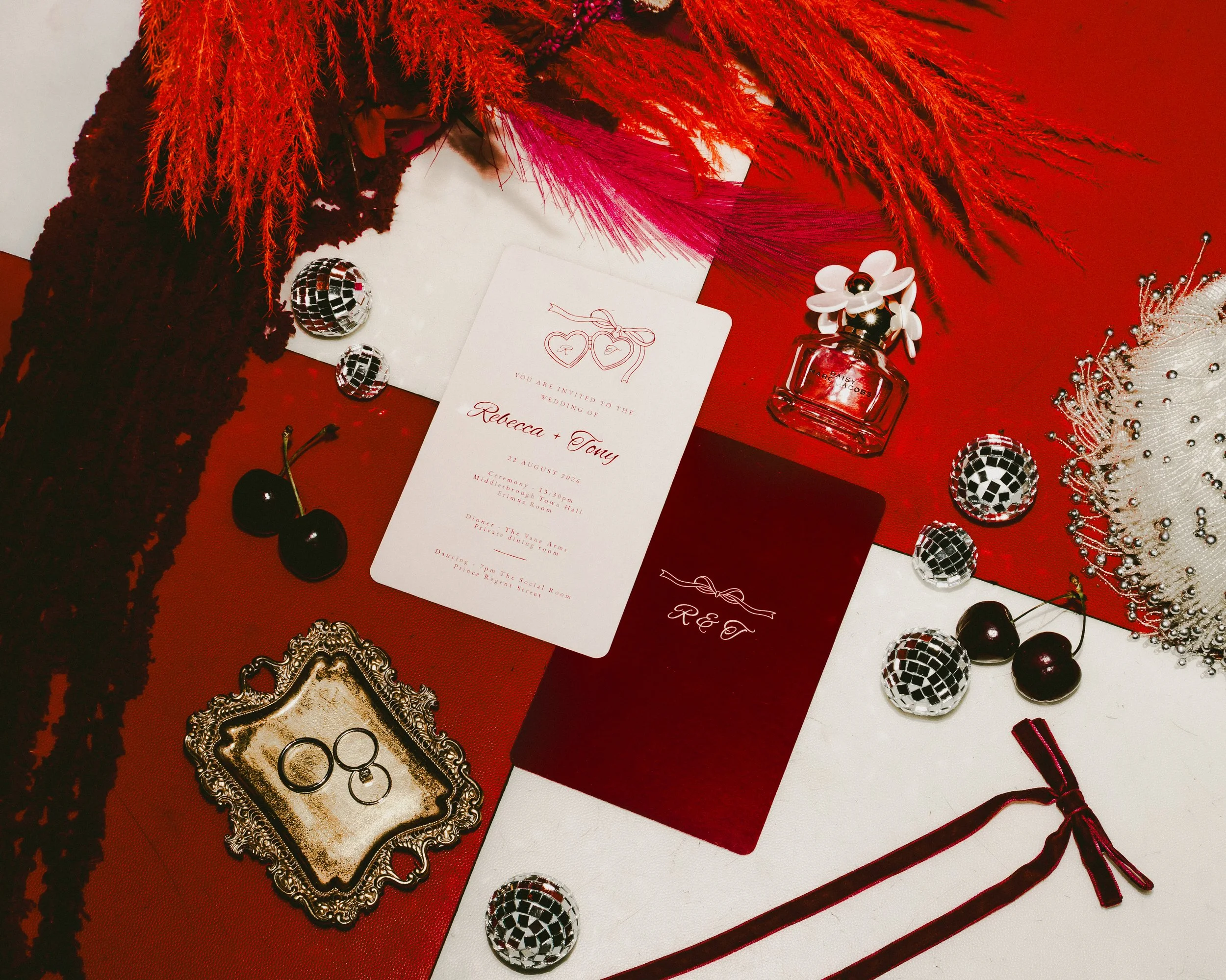 A wedding invitation surrounded by jewelry, perfume, and decorative accessories on a red and white surface.