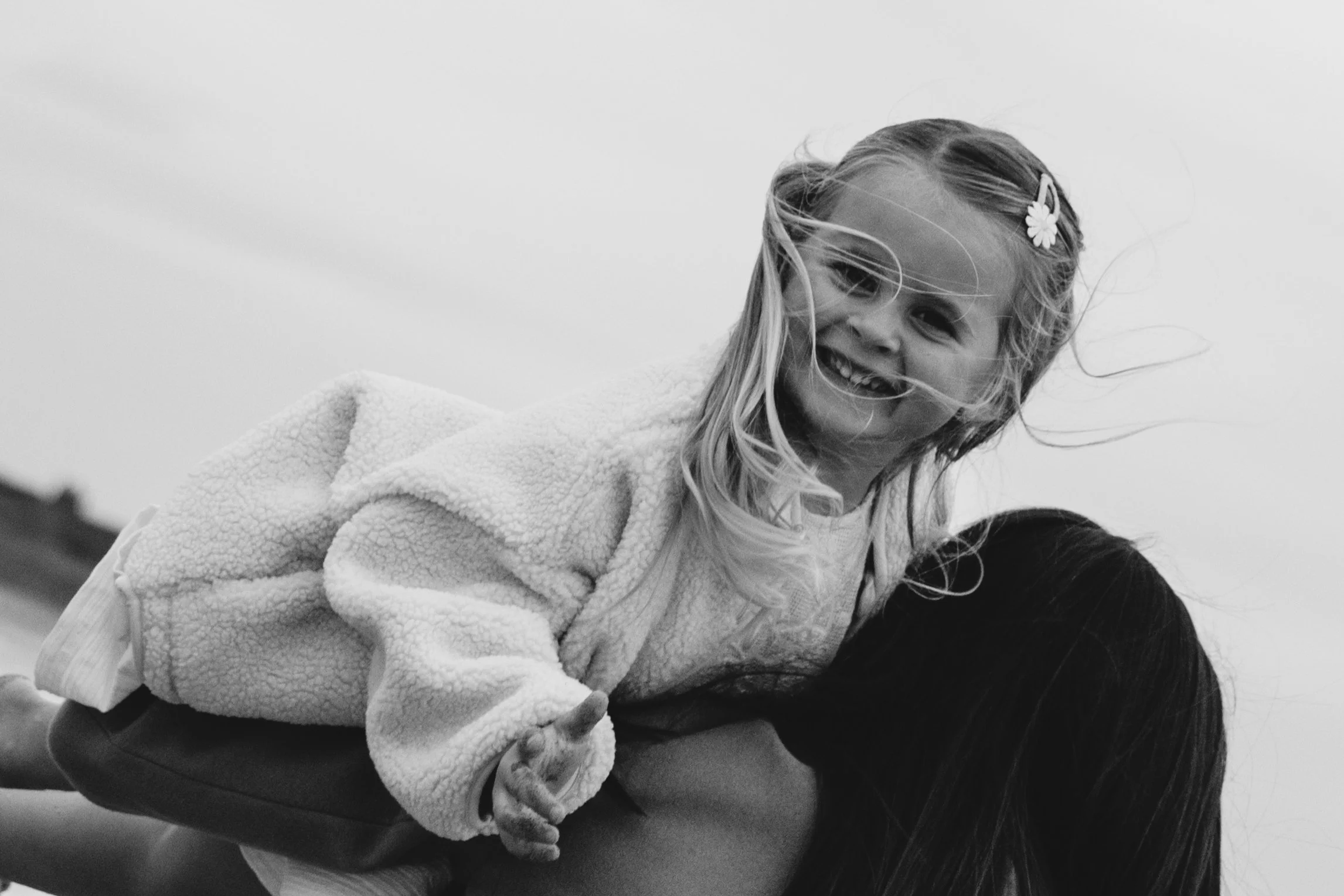 A young girl with long hair and a flower hair clip smiling and playing on the shoulders of a woman, both outdoors against a cloudy sky.