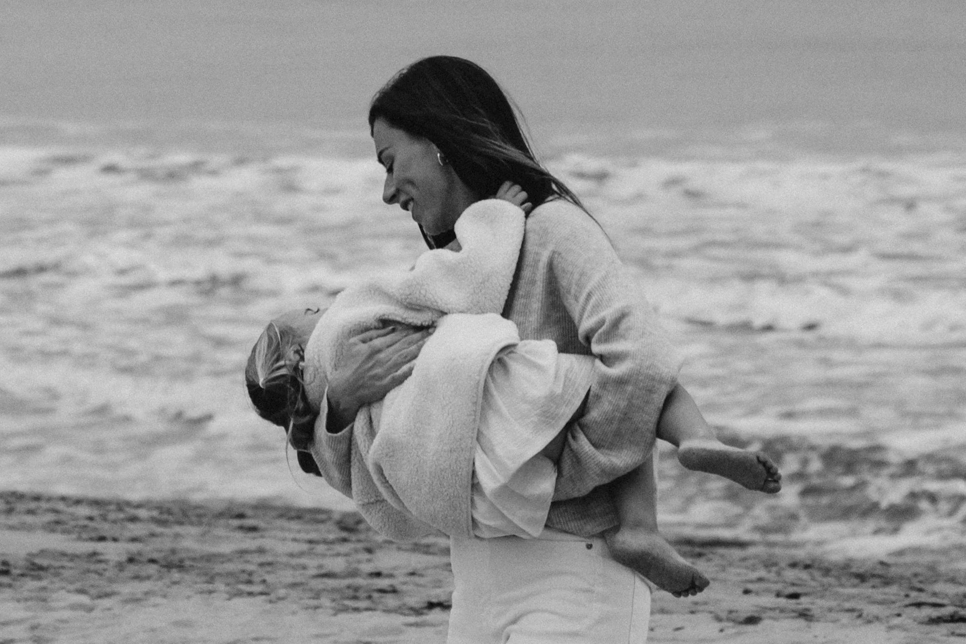 A woman is holding a child in her arms at the beach. They are smiling, and the woman appears to be joyful. The photo is in black and white with the beach and the ocean in the background.