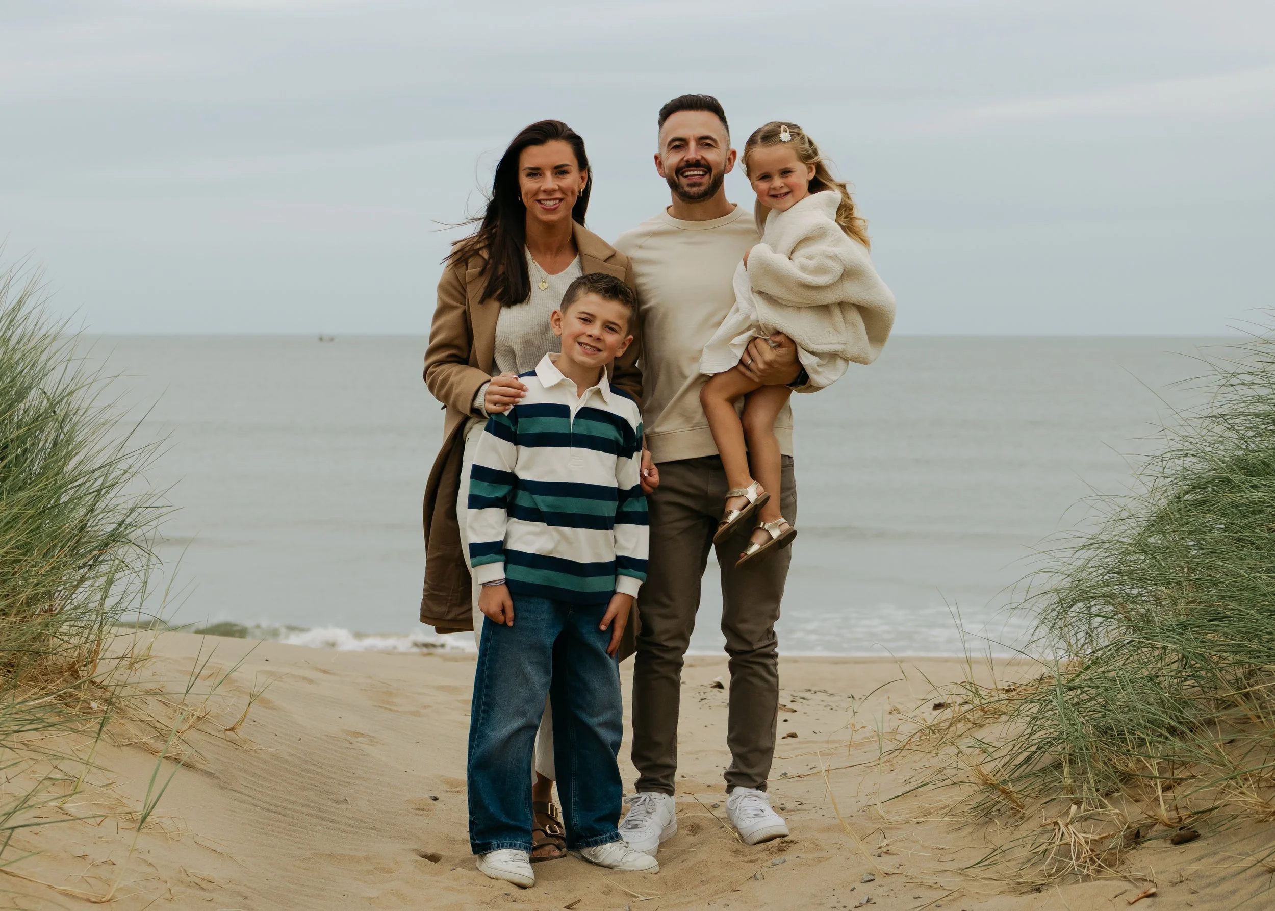 A family of four standing on a sandy beach with ocean in the background. The man is holding a young girl, the woman is smiling, and a boy is standing in front.