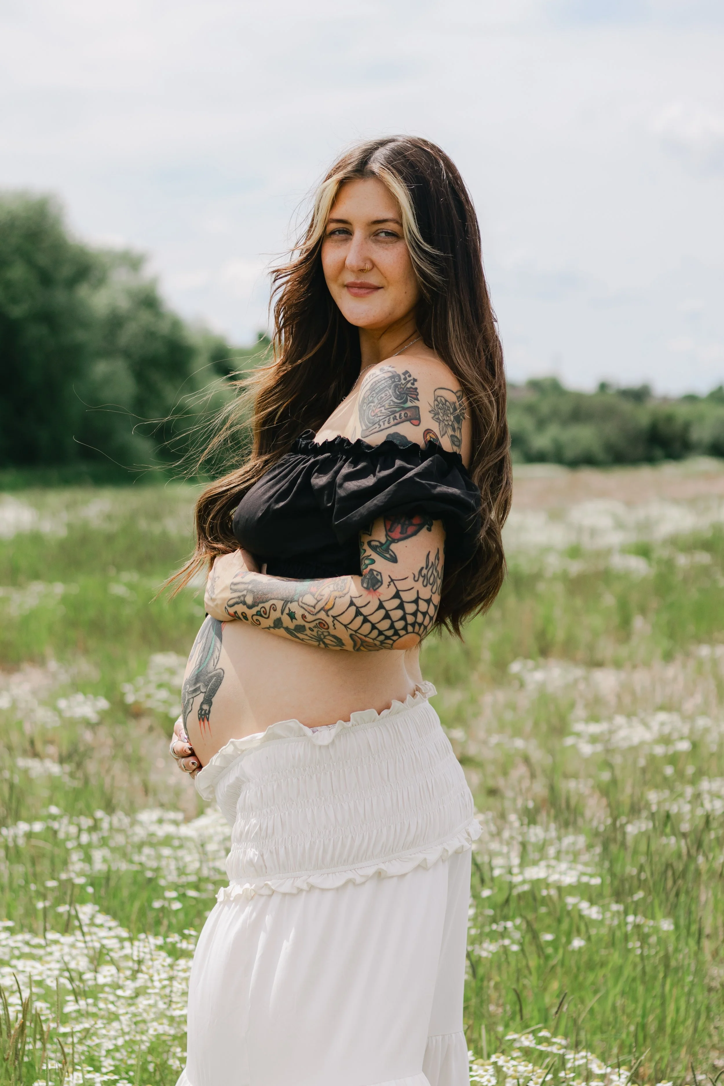 A woman with long dark hair and tattoos on her arms and shoulder standing outdoors in a grassy field with trees in the background, wearing a black off-the-shoulder top and white skirt.