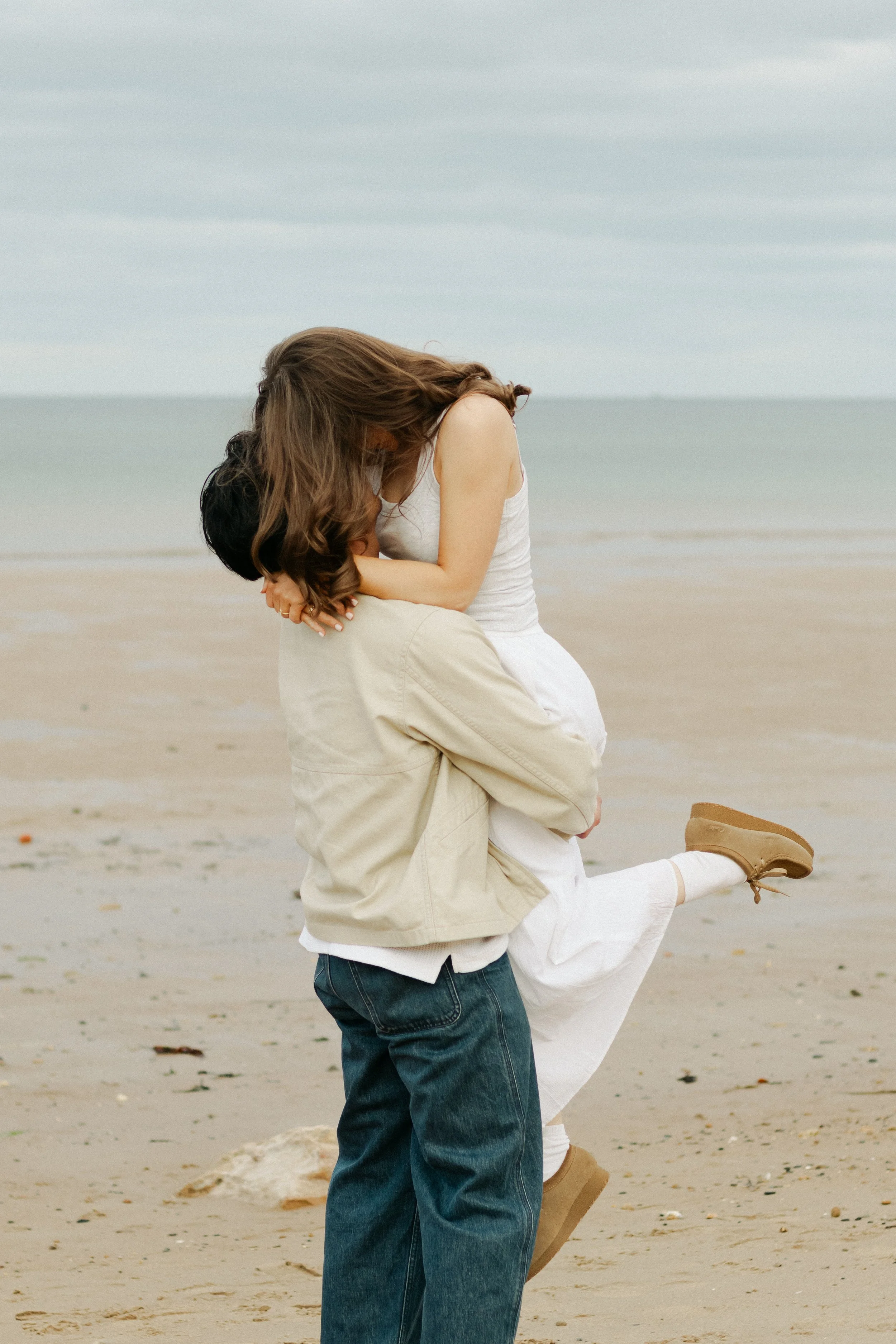 A couple at the beach, with the woman sitting on the man's lap, both with their heads bowed together, the woman leaning over the man's shoulder, surrounded by sand and ocean in the background.